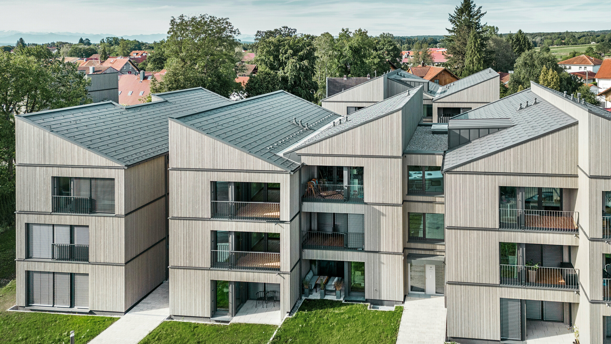 Vue aérienne de l'immeuble d'appartements moderne du quartier Schmucker à Utting am Ammersee, avec une construction hybride en bois durable et d'élégantes façades en bois. Les bâtiments sont équipés de R.16 PREFA durables en gris sombre P.10. Cette architecture innovante, conçue par WWA Architects de Munich, se caractérise par des lignes claires et une intégration harmonieuse dans l'environnement verdoyant. Les balcons spacieux et les grandes fenêtres assurent une atmosphère de vie lumineuse et conviviale, tandis que les matériaux de haute qualité garantissent durabilité et résistance aux intempéries.