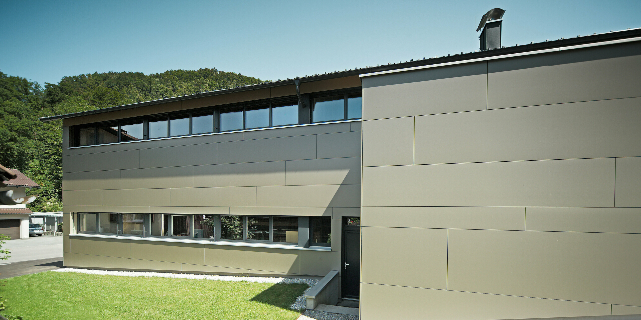 Ansicht eines Einfamilienhauses von der Gartenseite in Hohenems, verkleidet mit PREFA Aluminium Verbundplatten in der Farbe Bronzemetallic. Die harmonische Verbindung von moderner Architektur und Natur wird durch die große Fensterfront und die klaren Linien der Fassadenplatten unterstrichen, während die PREFA Dachplatten in P.10 Anthrazit für eine optische Einheit sorgen und das Haus vor den Elementen schützen.