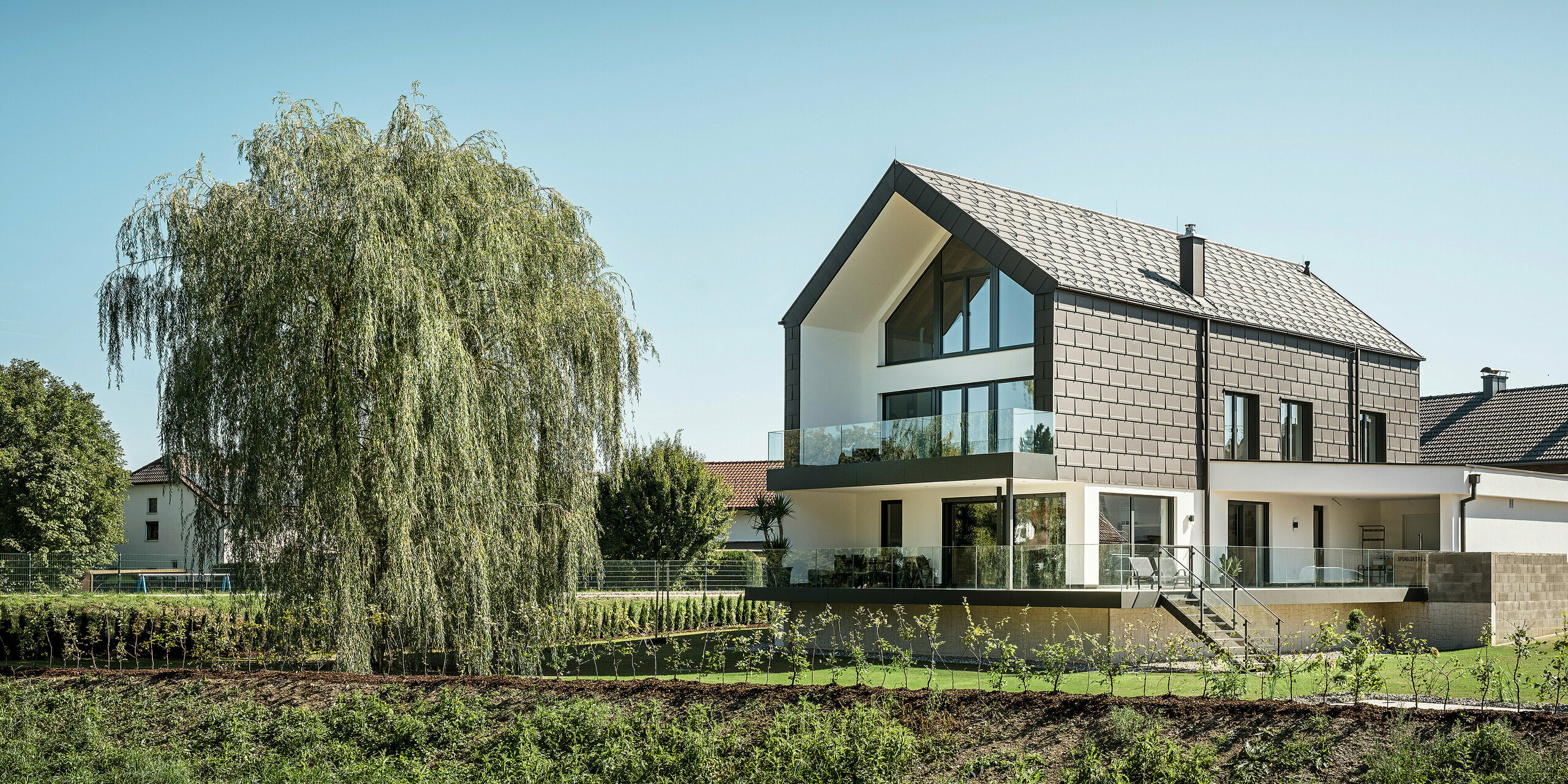 Einfamilienhaus in Neukirchen, Österreich, inmitten eines liebevoll angelegten Gartens. Die PREFA Dachplatte R.16 in braun P.10 fügt sich perfekt in das moderne Design des Hauses ein und schafft ein ästhetisches Zusammenspiel von traditionellen und modernen Stilelementen. Die großen Fenster öffnen das Gebäude zur umgebenden Natur und ermöglichen ein lichtdurchflutetes Wohnen. Die ruhige Wohnlage und die Einbindung der Gebäude in die natürliche Landschaft spiegeln die Ansprüche der Bauherren an die Lebensqualität wider.