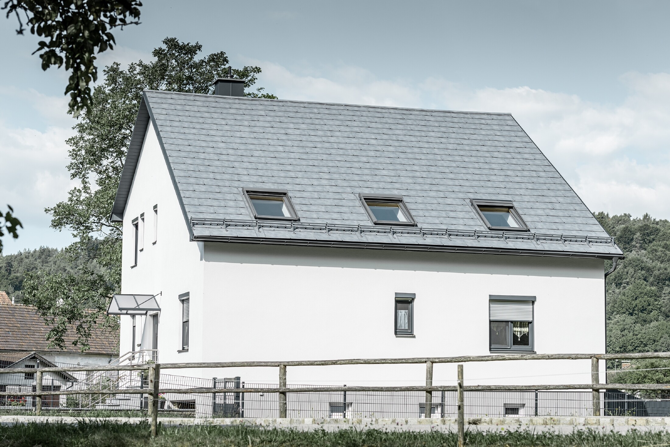 Le toit à deux pans de la maison individuelle classique a été recouvert avec la nouvelle tuile R.16 PREFA couleur gris pierre. Trois fenêtres de toit ont été intégrées dans la surface de toit et des arrêts neige ont été montés. La façade a été simplement laissée blanche.