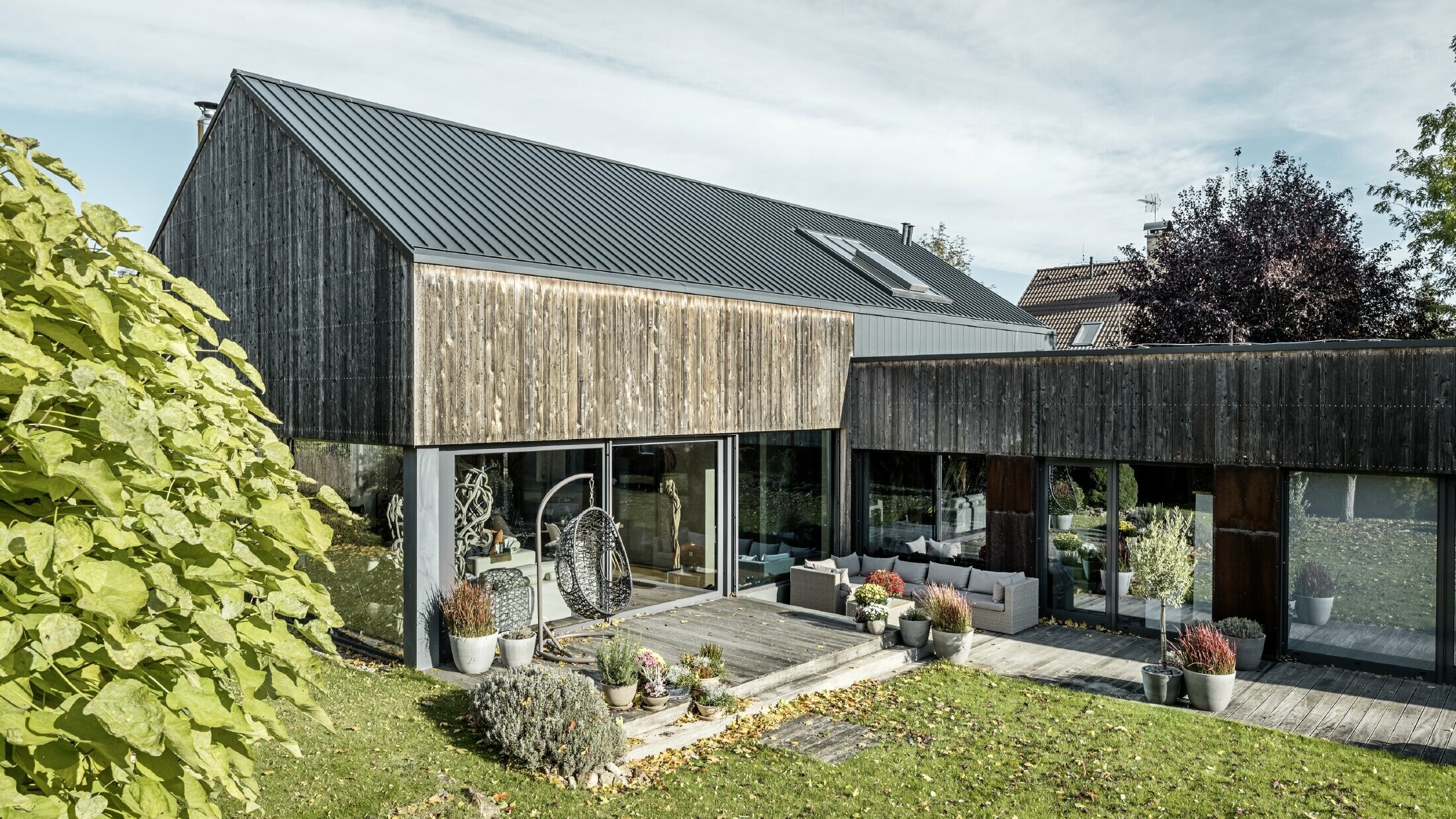 Maison individuelle avec toit à pignon, recouvert avec les joints debout double agrafe PREFALZ en anthracite et façade en bois patiné. Avec belle terrasse en bois et grandes fenêtres au rez-de-chaussée.