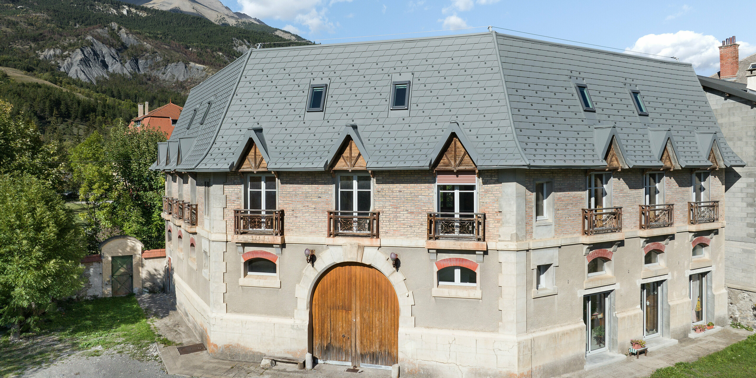 Frontansicht eines Anwesens in Barcelonnette, Frankreich. Das großflächige Dach wurde mit der PREFA Dachschindel in P.10 Hellgrau saniert. 