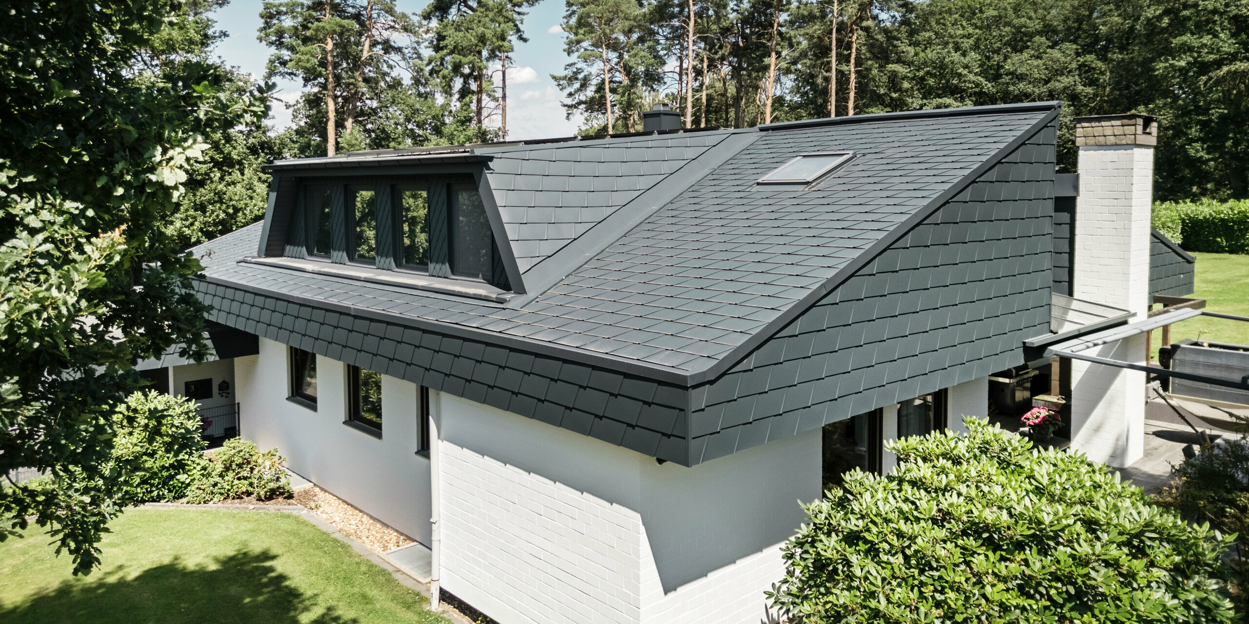 Vue aérienne d'une maison individuelle à Mechernich, en Allemagne, avec des bardeaux de toit DS.19 PREFA et une lucarne trapézoïdale revêtue en P.10 anthracite. Le toit est recouvert de bardeaux en aluminium durables, offrant une solution résistante aux intempéries et esthétique. La façade blanche de la maison s'harmonise avec la toiture sombre, tandis que les arbres et les buissons complètent l'environnement naturel de la maison.