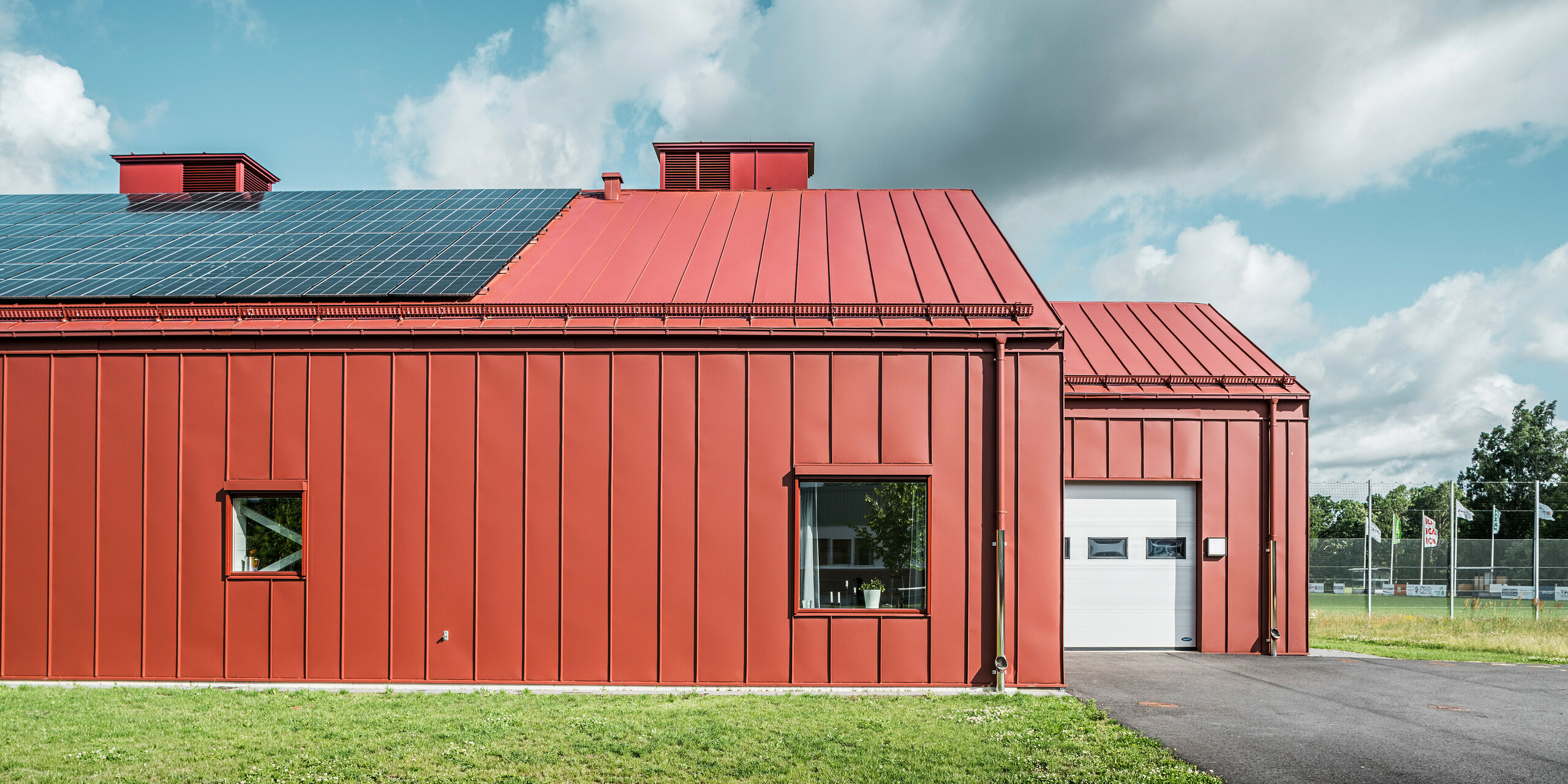 Vue latérale du restaurant Gröna Hjärtat, la cantine scolaire de Göingeskolan et Prästavångskolan à Broby, en Suède. La perspective montre la façade frappante et le toit avec revêtement à joints debout en PREFALZ rouge oxyde. Un système photovoltaïque à grande échelle sur le toit produit de l'électricité à partir d'énergies renouvelables. Le revêtement en aluminium durable de couleur rouge oxyde souligne le design moderne et économe en énergie du bâtiment. La gouttière PREFA s'intègre harmonieusement à la façade et s'adapte à l'aspect général du bâtiment.