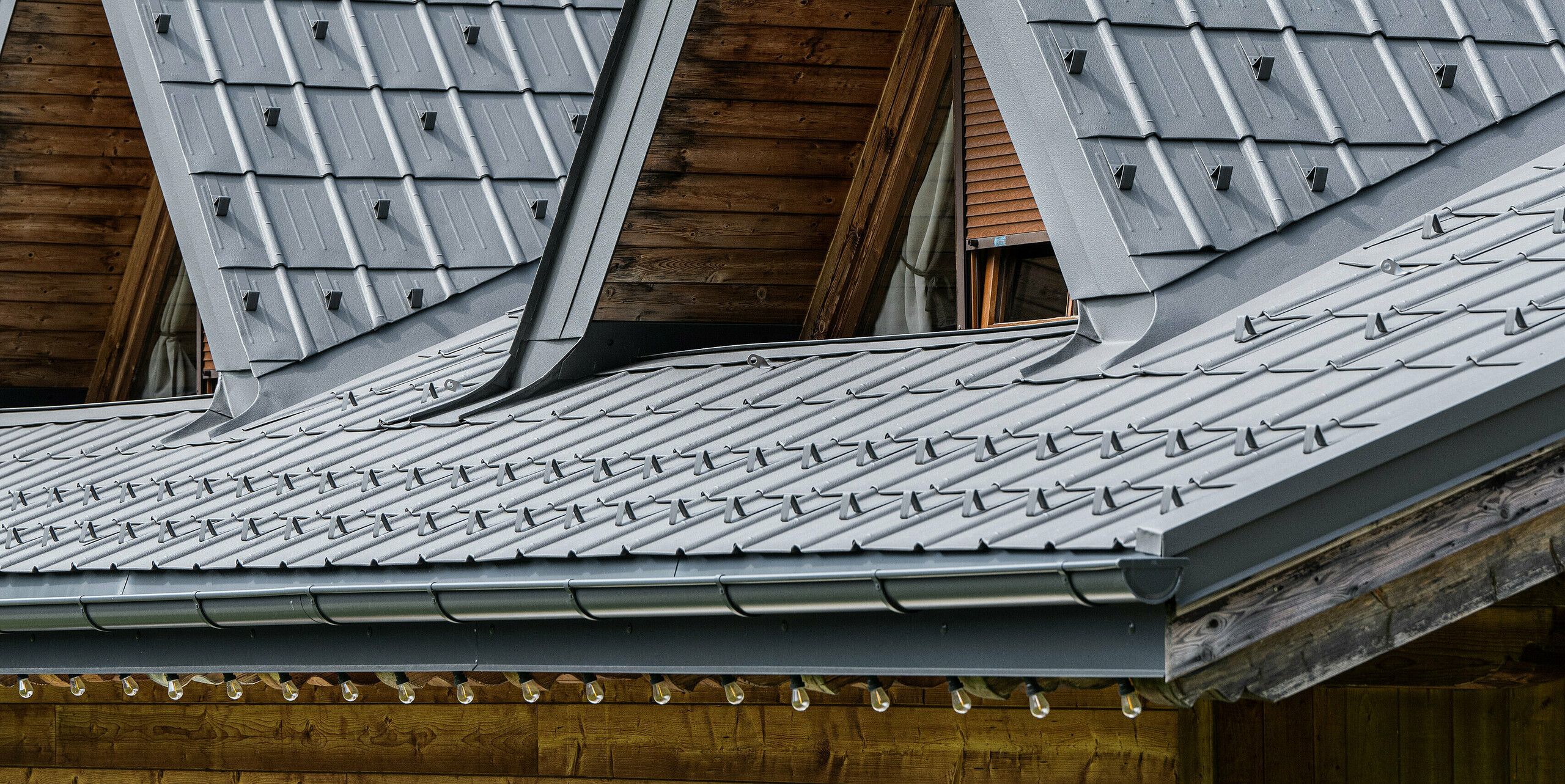 Detailní záběr typického alpského domu v Auronzo di Cadore se střešním systémem PREFA. Střecha se špičatými vikýři byla pokryta robustními falcovanými taškami. Hliníkové tašky PREFA odrážejí vysoce kvalitní a odolnou konstrukci, která zapadá do zalesněného horského pozadí. Střecha ukazuje dokonalou harmonii tradiční alpské dřevostavby a současné střešní krytiny, která je funkční i vzhledově působivá.
