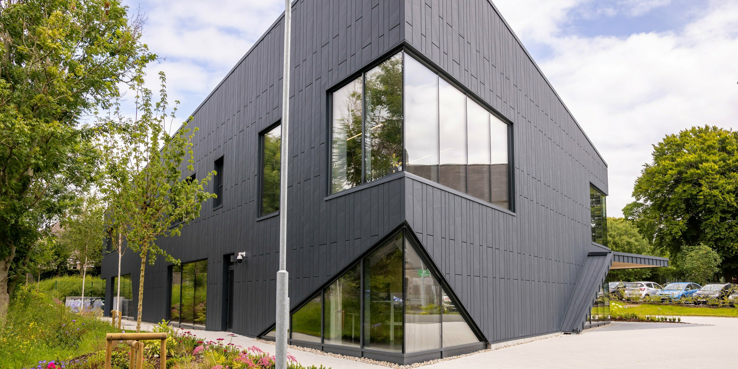 Façade du bâtiment CREW à Galway, Irlande, avec panneaux de façade PREFA Siding.X de couleur P.10 anthracite. Les lignes claires et les grandes surfaces vitrées soulignent l'architecture contemporaine du bâtiment. La façade en aluminium robuste et durable confère au bâtiment un aspect élégant et moderne, harmonieusement complété par la verdure environnante.