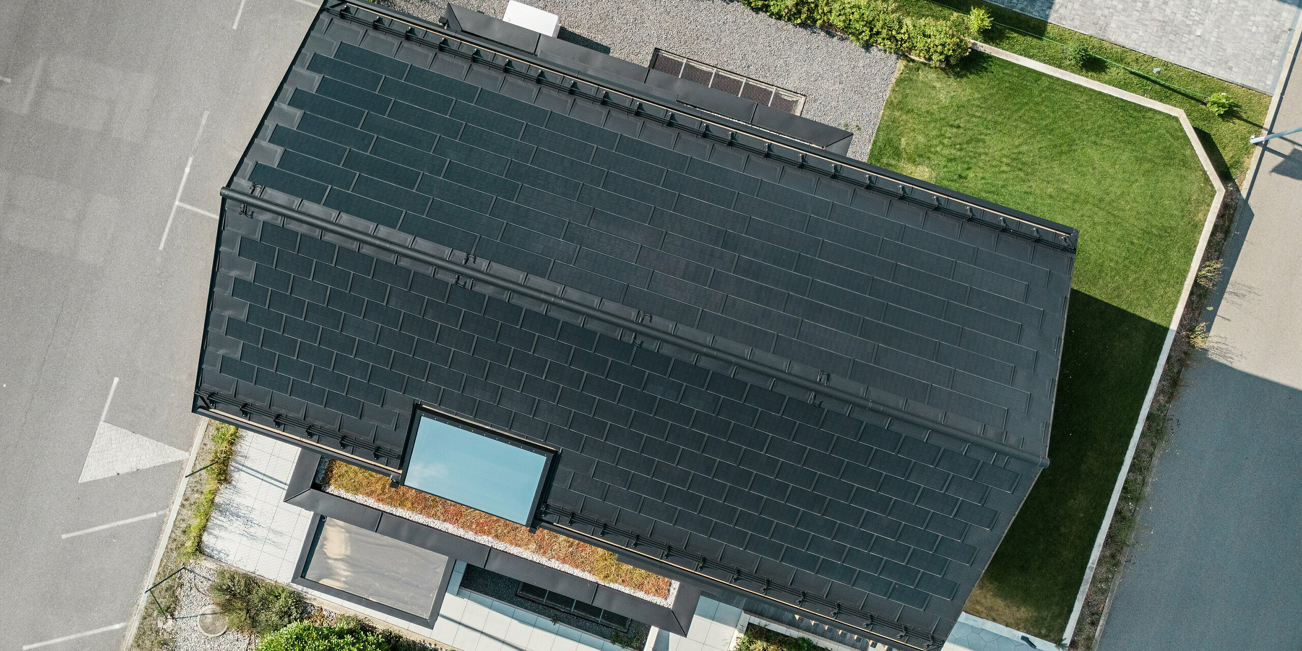 Vue aérienne de la maison témoin Autark dans le centre de maisons préfabriquées de Blaue Lagune, Vösendorf (AT), avec une toiture robuste en aluminium PREFA. La toiture est recouverte de tuiles solaires PREFA et de R.16 en P.10 noir. Le système photovoltaïque intégré garantit une production d'énergie durable en utilisant l'énergie du soleil. Le bardage en bois côté pignon complète l'aspect contemporain de la maison.