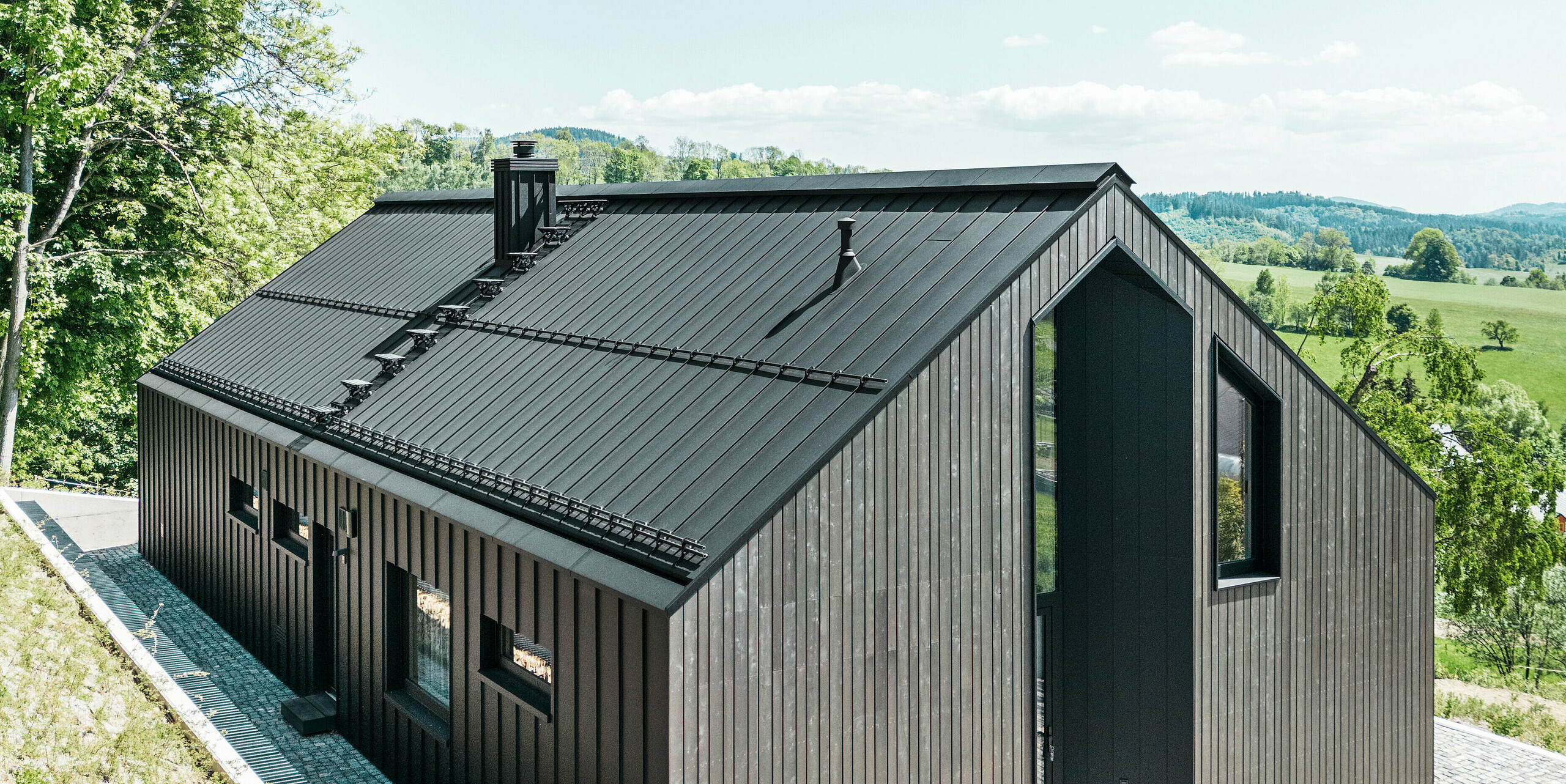 Dunkles Holzmassivhaus mit PREFALZ in P. 10 am Dach und der Rückseite des Gebäudes. Vor dem Haus steht ein kleines Nebengebäude mit einer Aluminiumhülle aus PREFALZ in P.10 Schwarz. Im Hintergrund ein strahlend blauer Himmel und Wald.