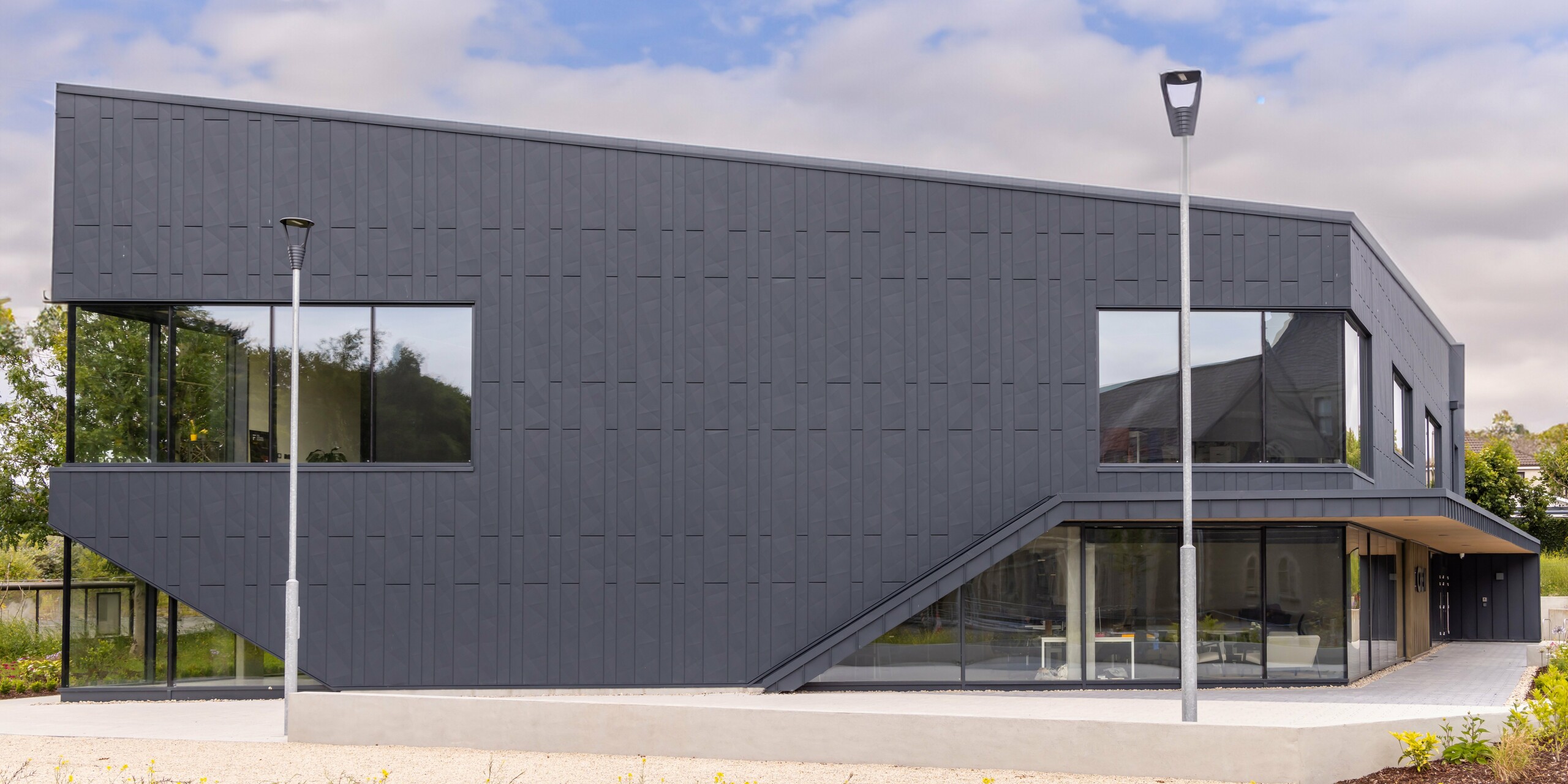 Vue latérale du bâtiment CREW à Galway, en Irlande, avec une façade moderne en aluminium PREFA composée de panneaux de façade Siding.X et PREFALZ en anthracite P.10. Les lignes géométriques et les grandes surfaces vitrées soulignent l'architecture contemporaine. Le revêtement en tôle de haute qualité confère au bâtiment un aspect élégant et professionnel, tandis que le paysage environnant complète le design moderne.