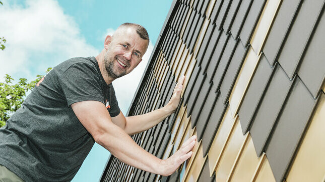 Artisan PREFA portant un t-shirt gris sur une façade en losanges PREFA couleurs brun et or maya.