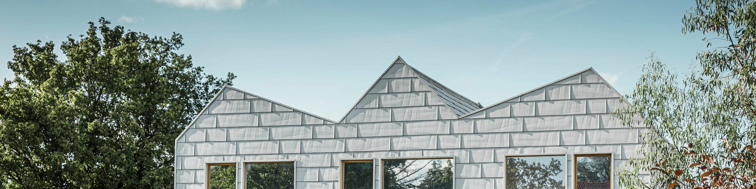 Modernes Doppelhaus mit Satteldach und PREFA-Fassade. Die Fassade besteht aus FX.12 Aluminiumpaneelen, die eine strukturierte, moderne Optik bieten. Das Haus ist von einem gepflegten Garten umgeben, und die Fensterrahmen harmonieren perfekt mit der Gesamtarchitektur.