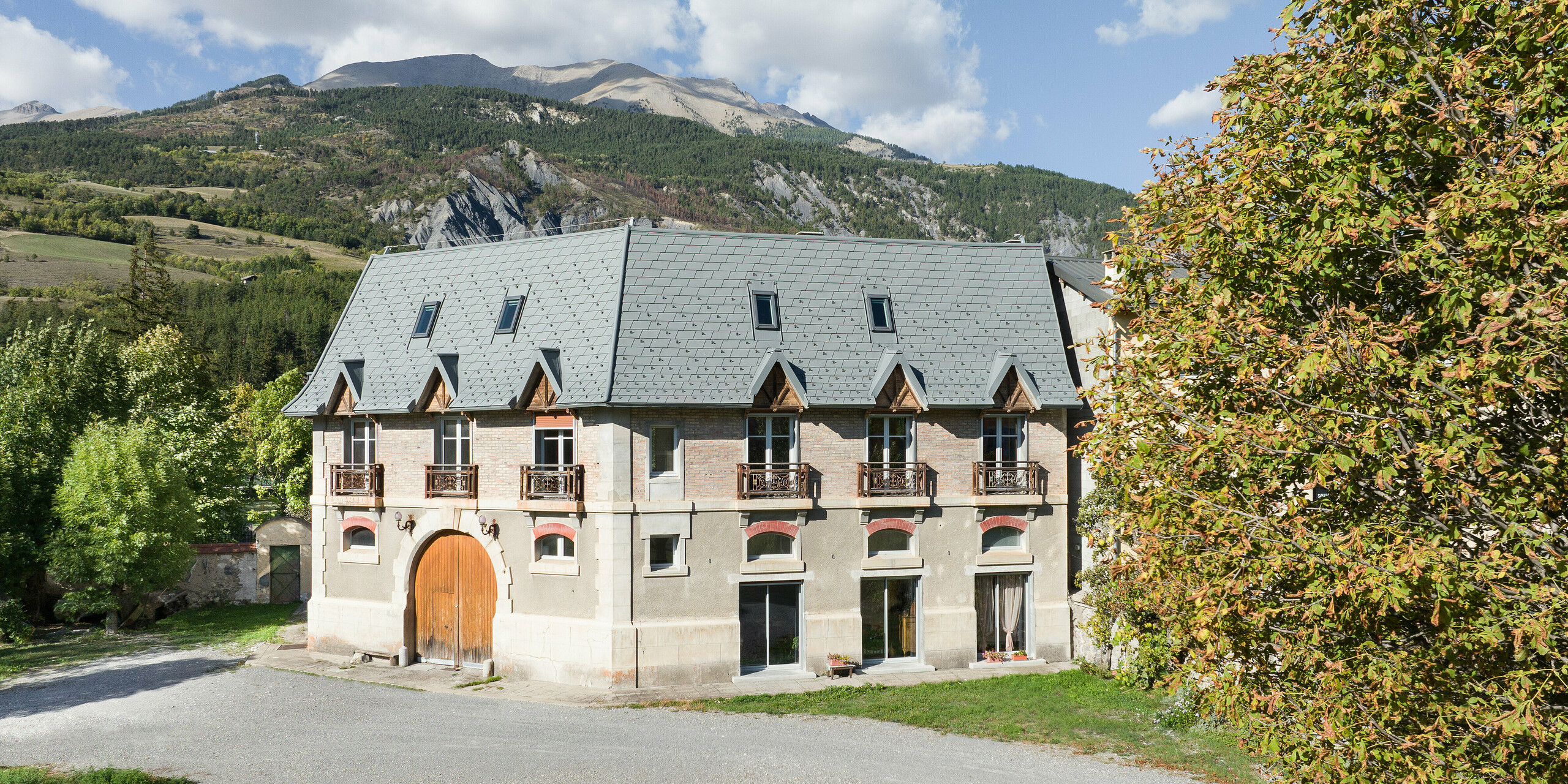 Frontansicht eines Anwesens in Barcelonnette, Frankreich. Das großflächige Dach wurde mit der PREFA Dachschindel in P.10 Hellgrau saniert. 