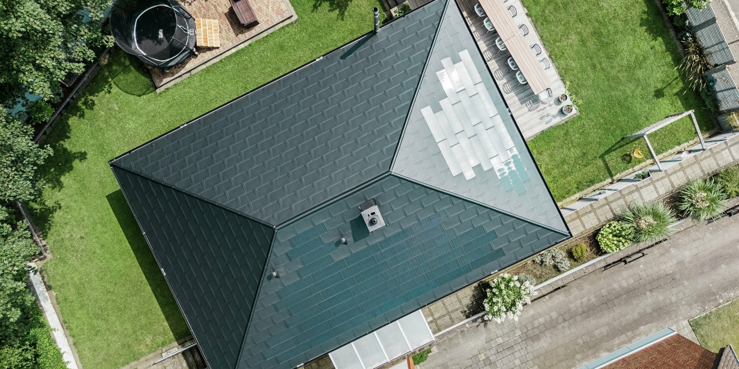 Vue plongeante sur un toit moderne en aluminium PREFA d'un bungalow à Rommerskirchen. La combinaison innovante de tuiles solaires et de R.16 en P.10 noir garantit une production d'énergie durable et une protection optimale. Le design élégant s'intègre parfaitement dans l'environnement verdoyant et crée une impression générale harmonieuse.