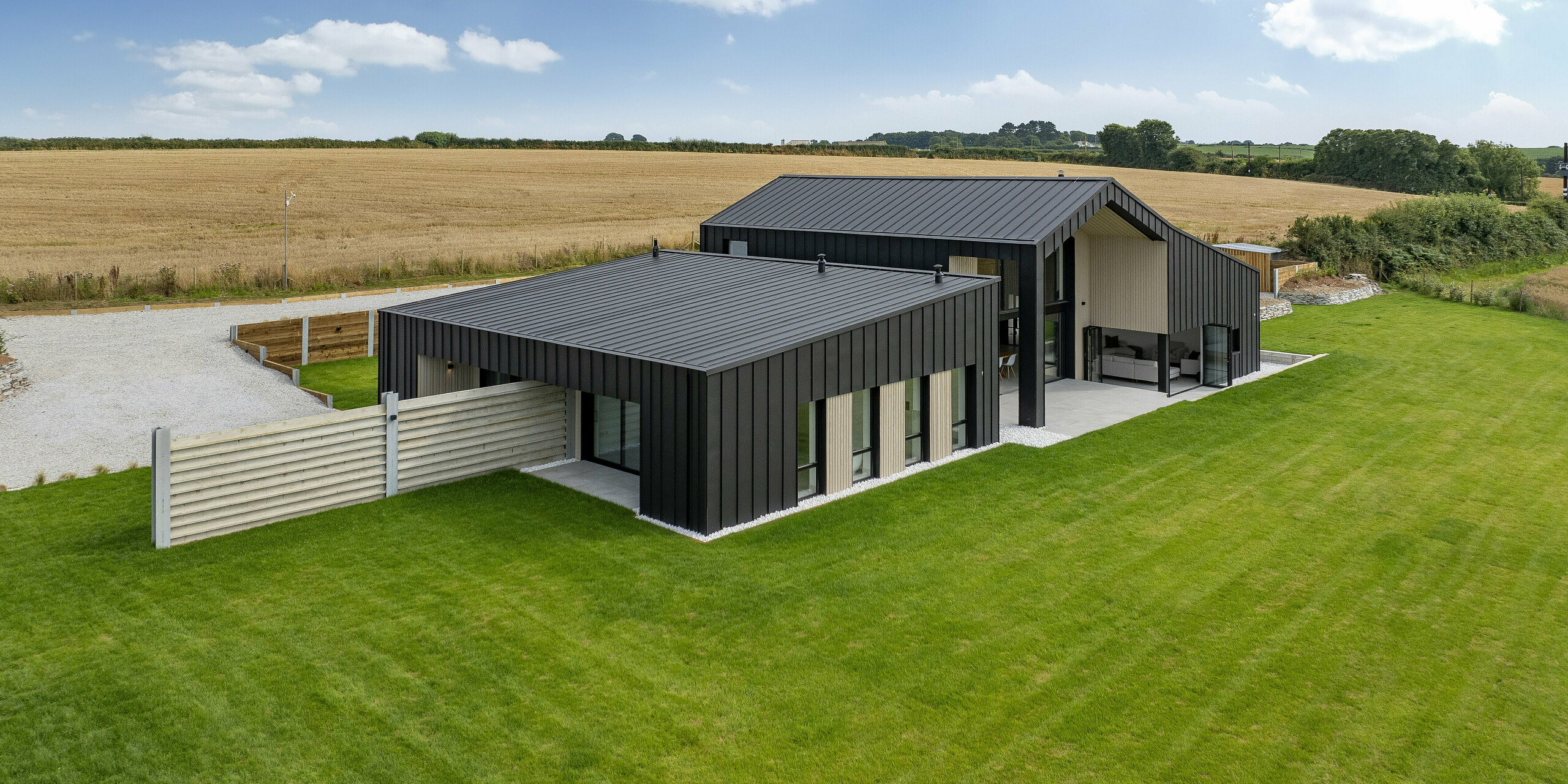 Vue grand angle de l'arrière de « The Hide » à Cornwall. La maison familiale moderne avec toit PREFALZ et revêtement de façade en aluminium s'intègre harmonieusement dans l'environnement rural avec de nombreux champs. La toiture à joint debout de couleur anthracite souligne le design minimaliste et offre en même temps une protection optimale contre les intempéries.