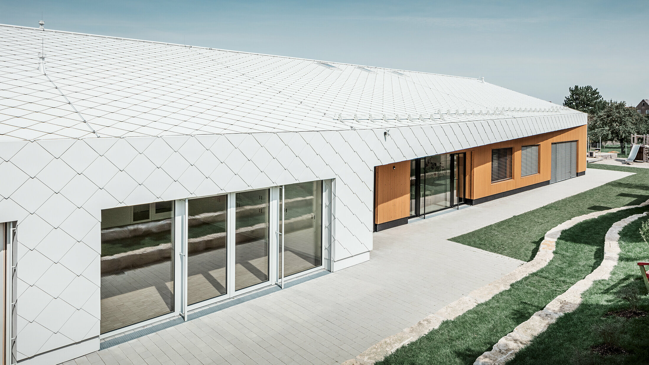 Jardin d’enfants revêtu du losange de toiture et de façade 44 de PREFA, en blanc Prefa ; la surface du toit et la façade ne font qu'un ; grandes surfaces vitrées