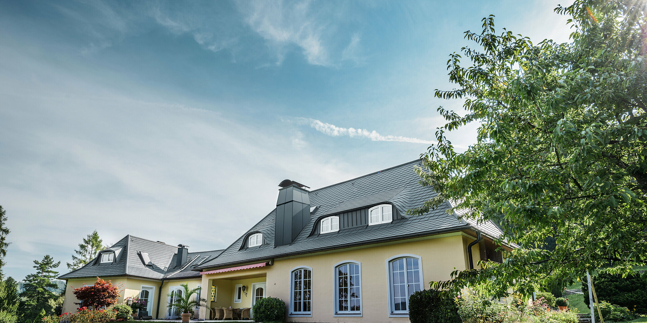 Maison individuelle classique avec lucarne arrondie, recouverte des bardeaux de toiture PREFA résistants aux tempêtes, avec garantie matériau de 40 ans.