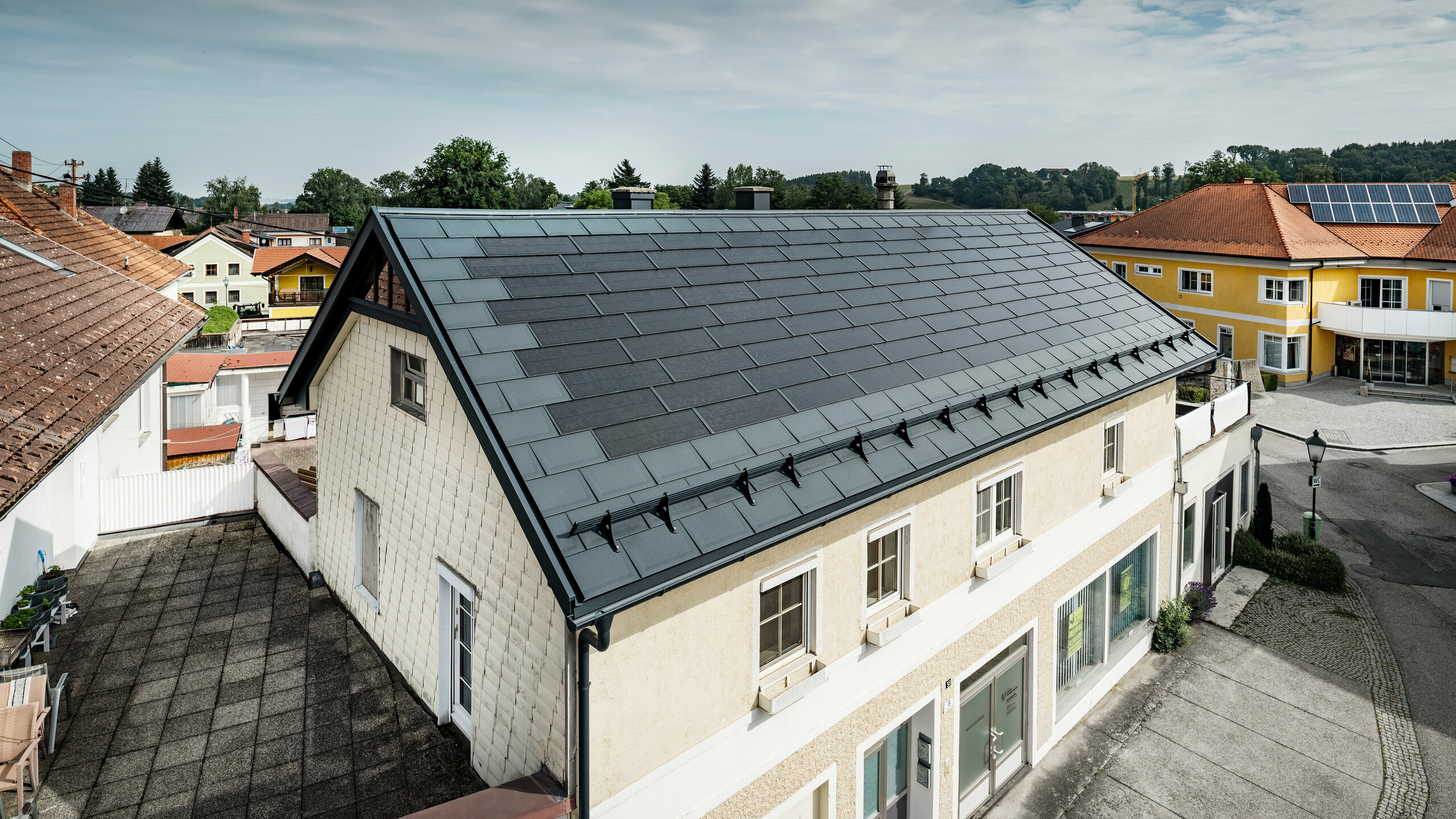 Vue du dessus d’une maison familiale à Mettmach, en Autriche. Le bâtiment le plus ancien a été rénové avec des tuiles solaires PREFA de couleur anthracite. Les systèmes de protection contre la neige et les lignes claires des panneaux solaires soulignent la finition soignée des produits en aluminium PREFA. Le toit de tuiles orange d’un immeuble voisin contraste en arrière-plan. Les modules photovoltaïques sont installés sur le toit de la maison individuelle voisine dans une solution de toit classique, qui montre la diversité des conceptions de toit économes en énergie.