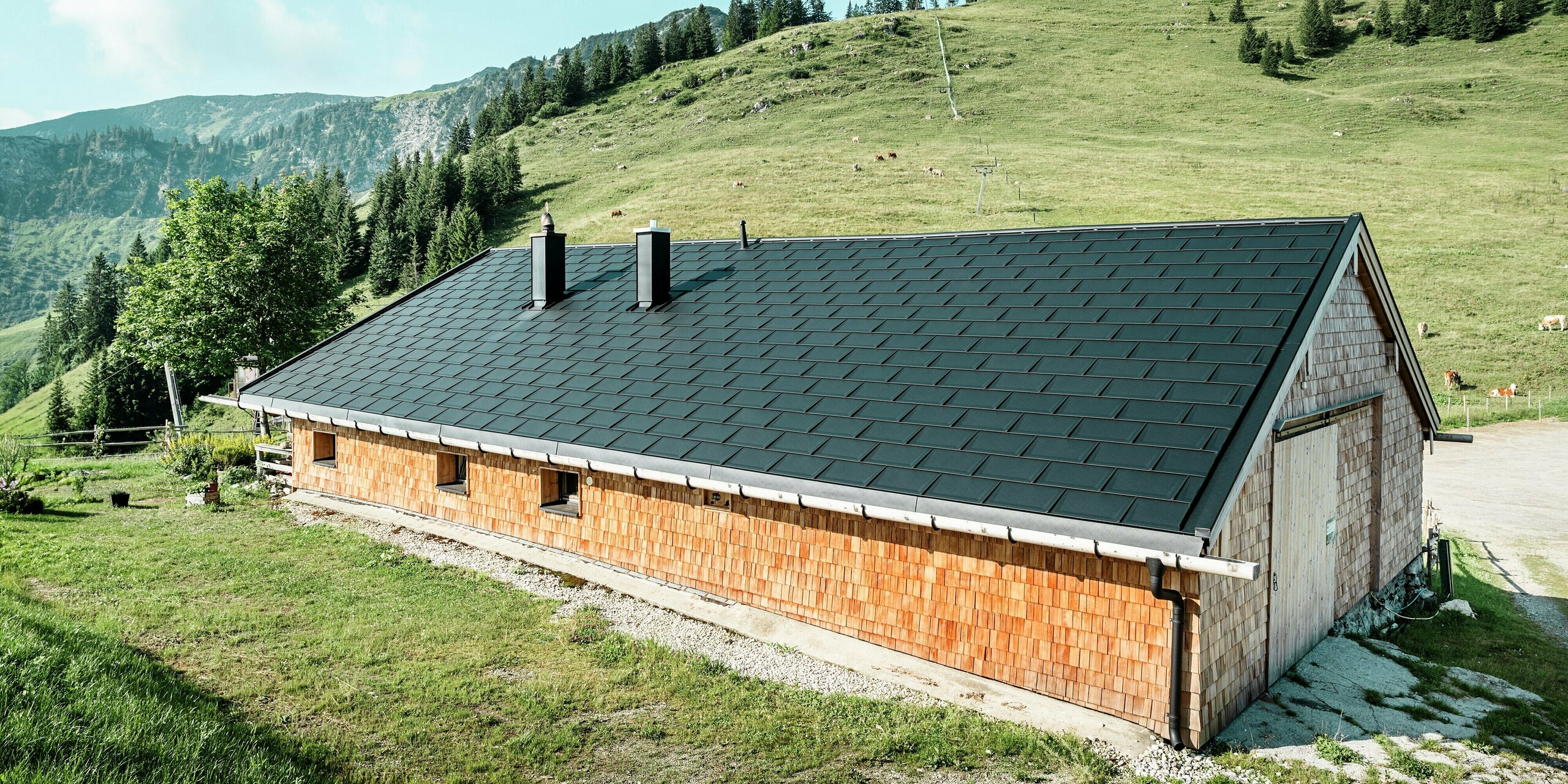 Rückansicht der Birkehütte auf der Walleralm in Bayrischzell, eingebettet in die majestätische Bergkulisse. Im Zuge einer umfassenden Sanierung wurde das Dach der Holzhütte mit robusten PREFA Dachplatten R.16 in der Farbe P.10 Schwarz eingedeckt. Auf der Sonnenseite des Daches wurden PREFA Solardachplatten verlegt, die für eine nachhaltige Energiegewinnung durch die Kraft der Sonne sorgen. Das hochwertige Blechdach schützt die Hütte vor den alpinen Witterungseinflüssen.