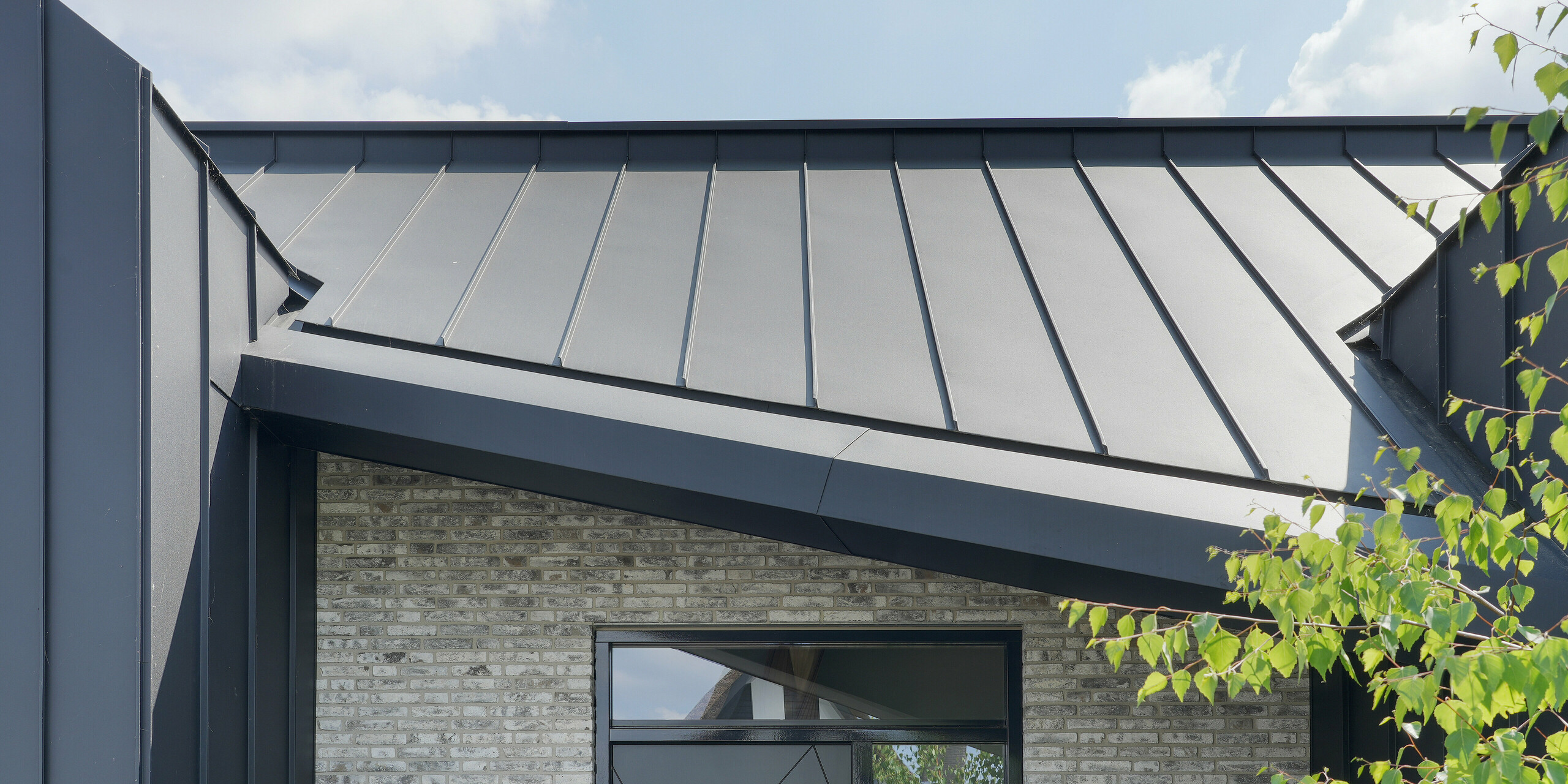 Entrée d'une élégante maison familiale à Veenendaal, Pays-Bas. L'entrée est mise en valeur par un revêtement en aluminium noir. Le système de toiture et de façade PREFALZ contraste avec la pierre naturelle gris clair de la façade. La grande porte noire porte le numéro de la maison 40 et offre un point de bienvenue élégant à côté du paysage de jardin aménagé de manière attrayante.