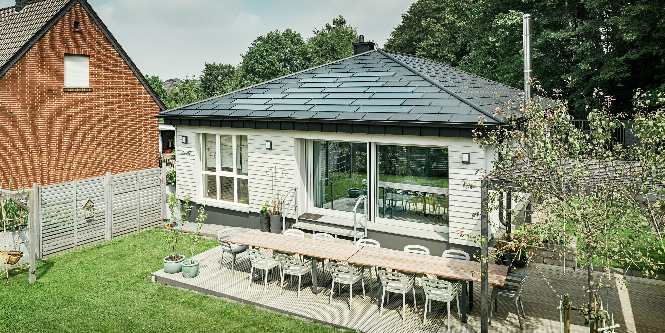 Bungalow rénové à Rommerskirchen avec une terrasse spacieuse, des fenêtres accueillantes et un toit en aluminium PREFA de haute qualité. Les tuiles solaires PREFA produisent de l'électricité durable et lesR.16 en P.10 noir offrent une protection optimale et une apparence élégante. Fonctionnalité et design moderne parfaitement combinés. Devant la maison individuelle se trouve un jardin bien entretenu avec un coin salon accueillant sur la terrasse.