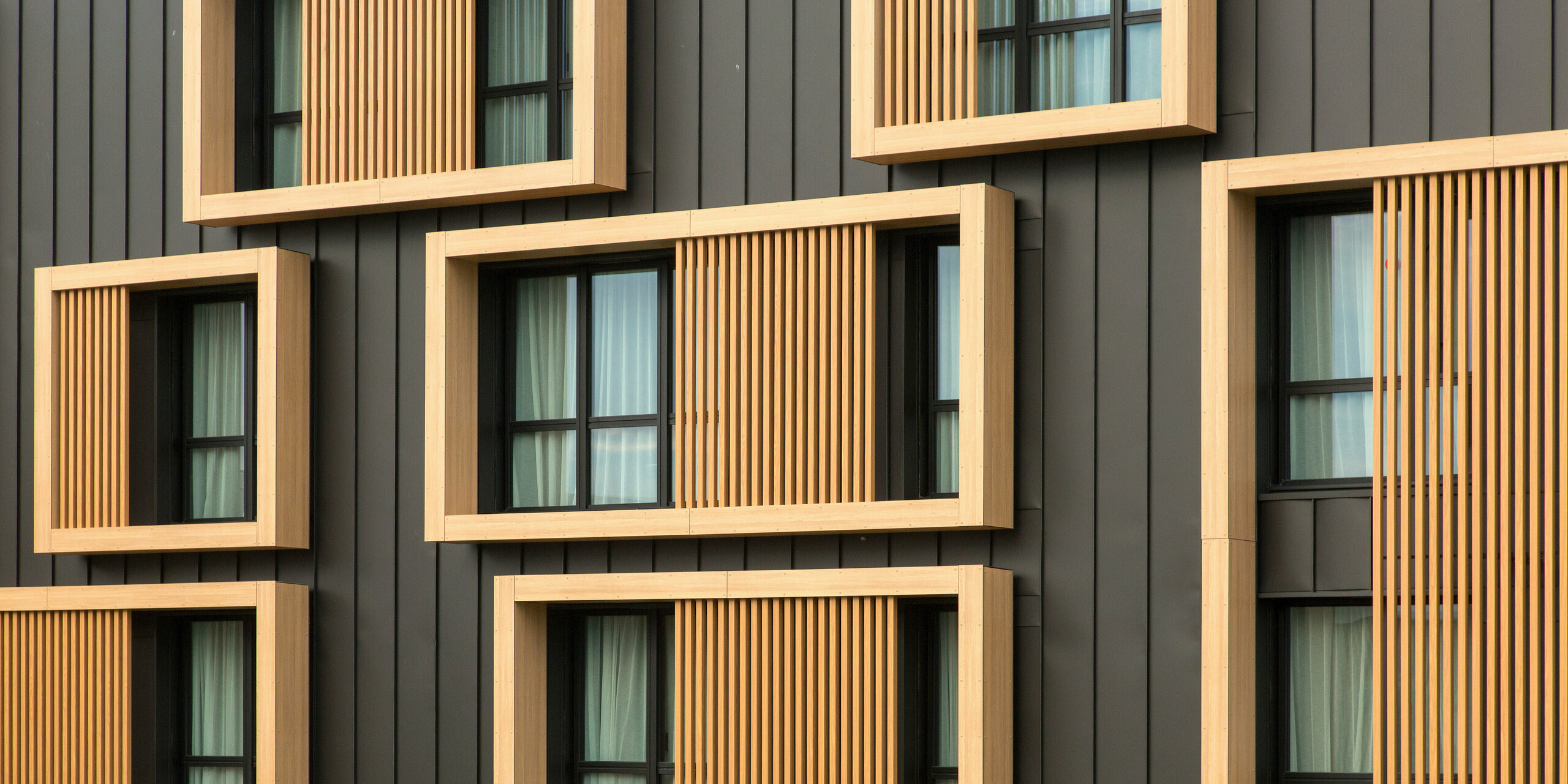 Gros plan de la façade de l'Hôtel Parc du Landreau aux Herbiers, France, avec une combinaison de PREFALZ gris noir et de cadres en bois autour des fenêtres. Le revêtement vertical en aluminium à joint debout est complété par les cadres de fenêtres en bois, qui ajoutent une touche chaleureuse et naturelle à l'architecture moderne.