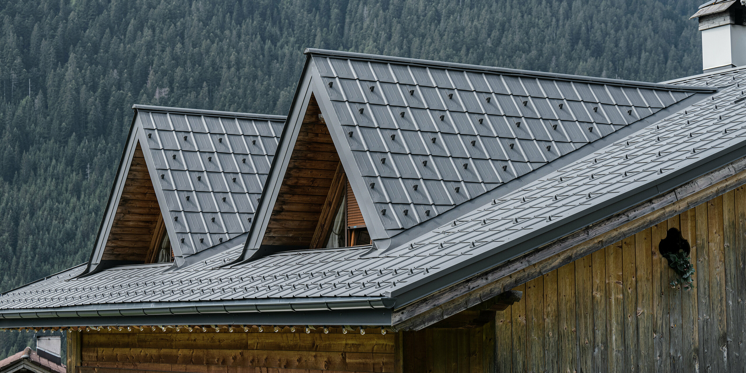 Detailní záběr na alpský dům v Auronzo di Cadore s nově instalovanou střechou PREFA v barvě P.10 světle šedé. Hliníkové falcované tašky nabízejí moderní dotek tradičního alpského architektonického stylu. Svou texturou a barvou vytvářejí hliníkové produkty harmonický vztah s okolní lesní krajinou. Tento obrázek ilustruje kombinaci regionálního řemesla s inovativními stavebními materiály s dlouhou životností.