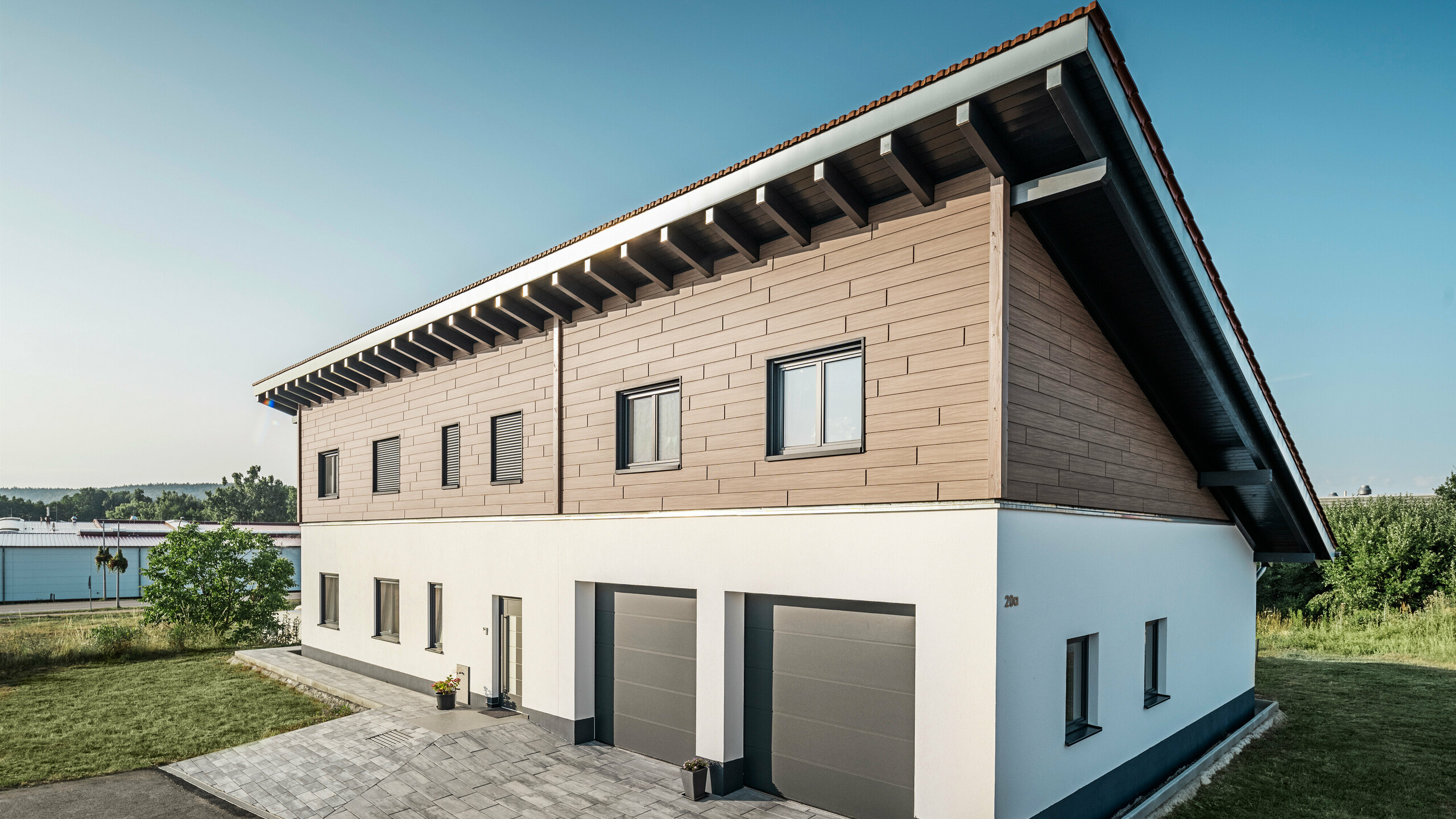 Vue latérale d'une maison individuelle avec un toit à une pente à Bruck. L'étage supérieur est enveloppé de Sidings PREFA en noyer foncé. Sur le côté droit de l'image, on peut voir deux portes de garage en anthracite. Au centre du bâtiment se trouve la porte d'entrée. Le socle est d'un blanc sobre. 