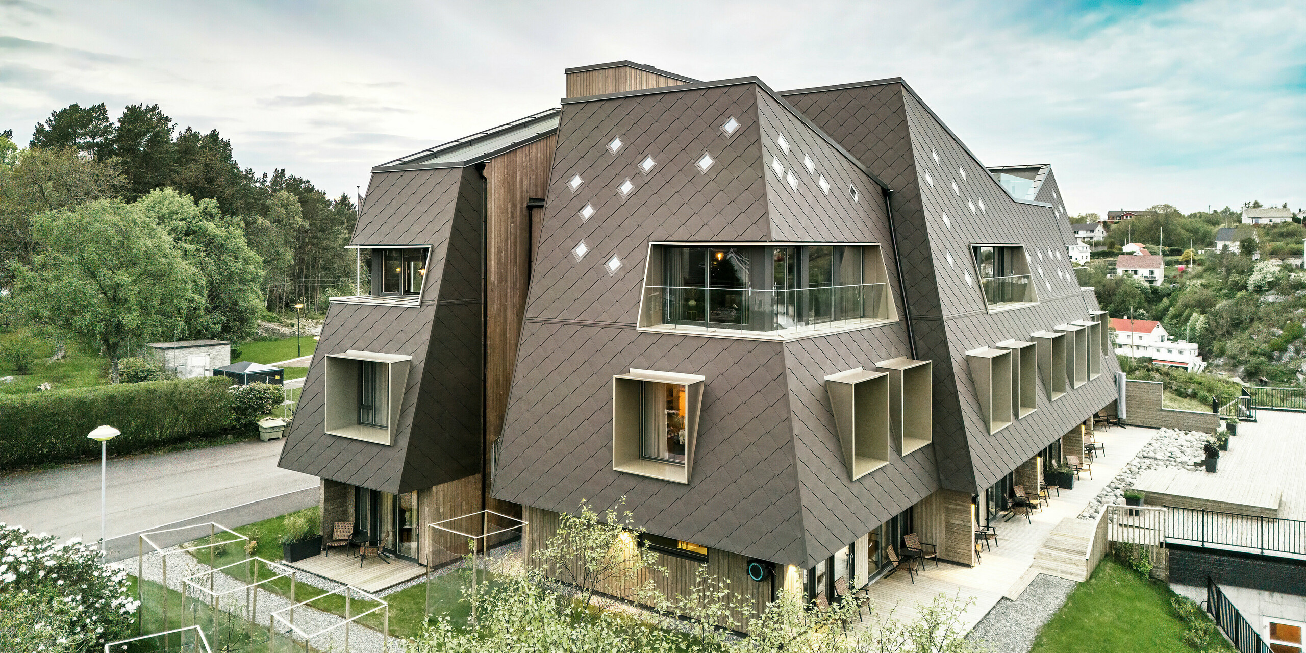 Façade de la maison Beckerwyc à Bekkjarvik, en Norvège, avec une façade saisissante composée de losanges PREFA 44 × 44 en brun noisette. L'image montre l'architecture futuriste du bâtiment avec des toits en pente et des éléments de fenêtres saillants. Les terrasses et balcons spacieux ainsi que le paysage verdoyant environnant soulignent l'interaction harmonieuse entre la nature et la construction moderne. L'enveloppe robuste en aluminium de haute qualité assure une protection durable.