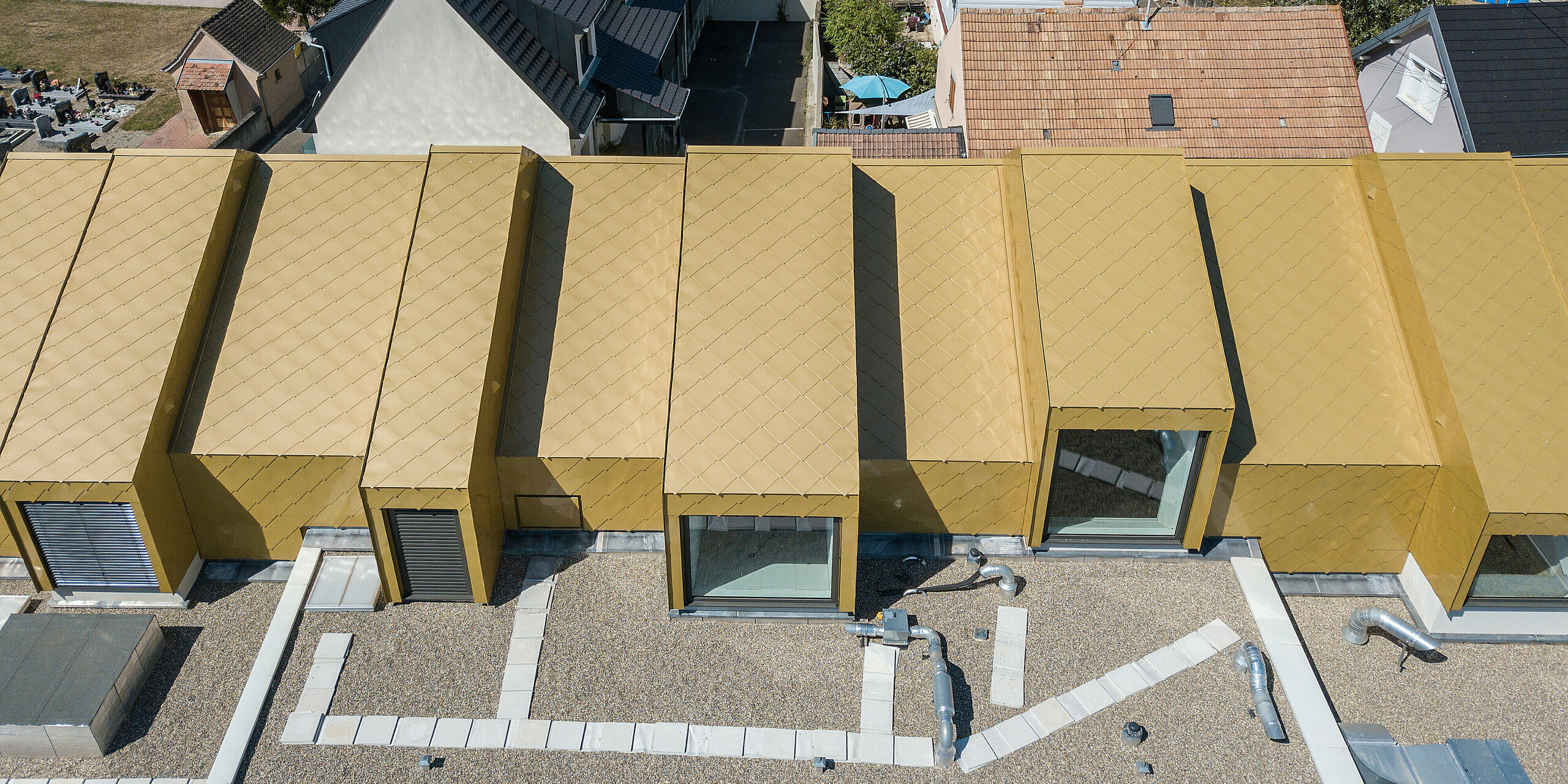 Vue plongeante sur le toit doré du centre communautaire et culturel de Fessenheim en France. Pour le projet de rénovation, des losanges de toiture et de façade PREFA 44 × 44 en Sparkling Gold ont été utilisés.