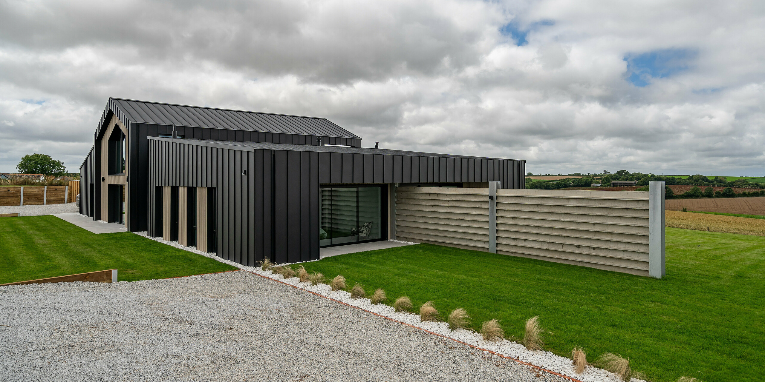 Vue latérale de « The Hide » en Cornouailles avec façade et toiture en aluminium PREFALZ en P.10 anthracite. L'aspect moderne des joints debout s'harmonise avec les lignes claires du bâtiment et les espaces naturels environnants. Le jardin devant et la terrasse abritée soulignent le caractère fonctionnel et élégant de la maison.