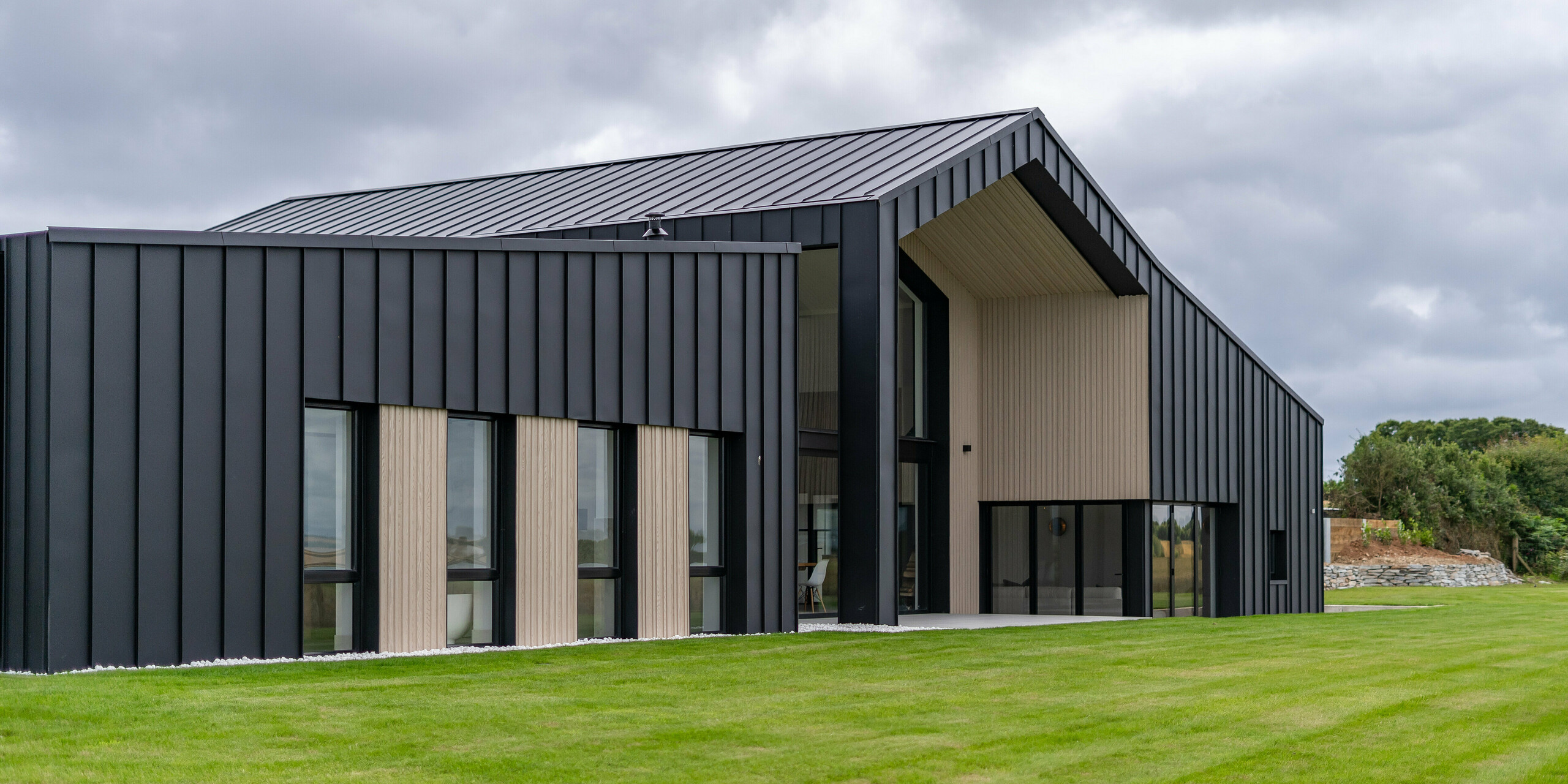 Vue oblique de l’arrière de la maison individuelle ‘The Hide’ à Cornwall, accentuée par la façade en aluminium PREFALZ en P.10 anthracite. Le toit à joint debout précis en tôle de haute qualité forme un contraste élégant avec les éléments en bois, tandis que le toit généreux avec ses lignes claires souligne l'architecture moderne du bâtiment. Entouré d'une verdure soignée, le projet s'intègre harmonieusement dans le paysage.