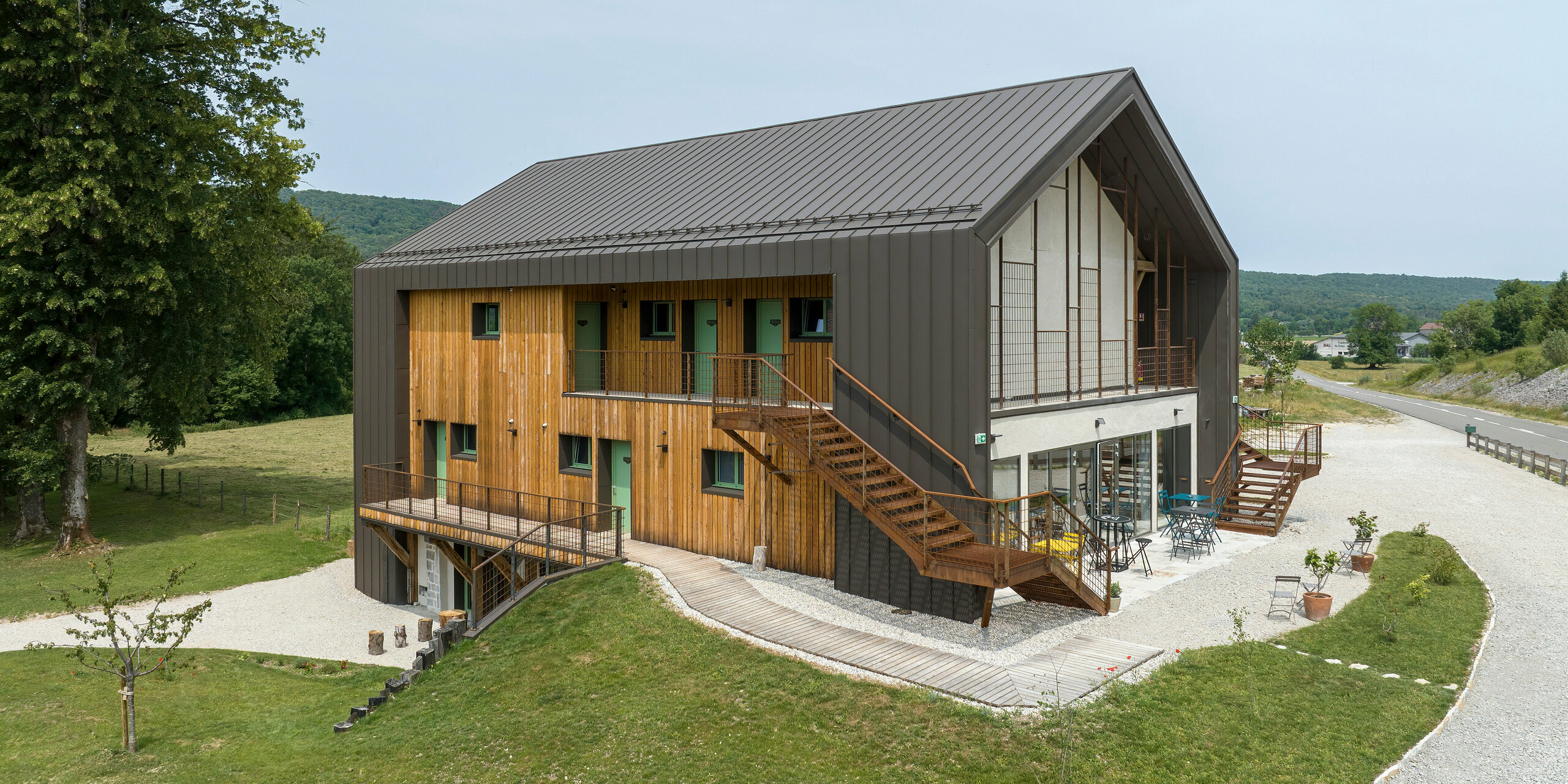 Bâtiment moderne avec toit en aluminium PREFA et façade en bois, également encadrée en aluminium PREFA. Les éléments en aluminium sont en PREFALZ de couleur P.10 brun. L'Hôtel en Tilleul à Pont du Navoy est entouré d'un paysage verdoyant. Les caractéristiques extérieures comprennent une allée de gravier, une pelouse paysagée et de jeunes arbres. Un escalier extérieur mène à l'étage supérieur, tandis qu'au rez-de-chaussée se trouve une terrasse avec des sièges.