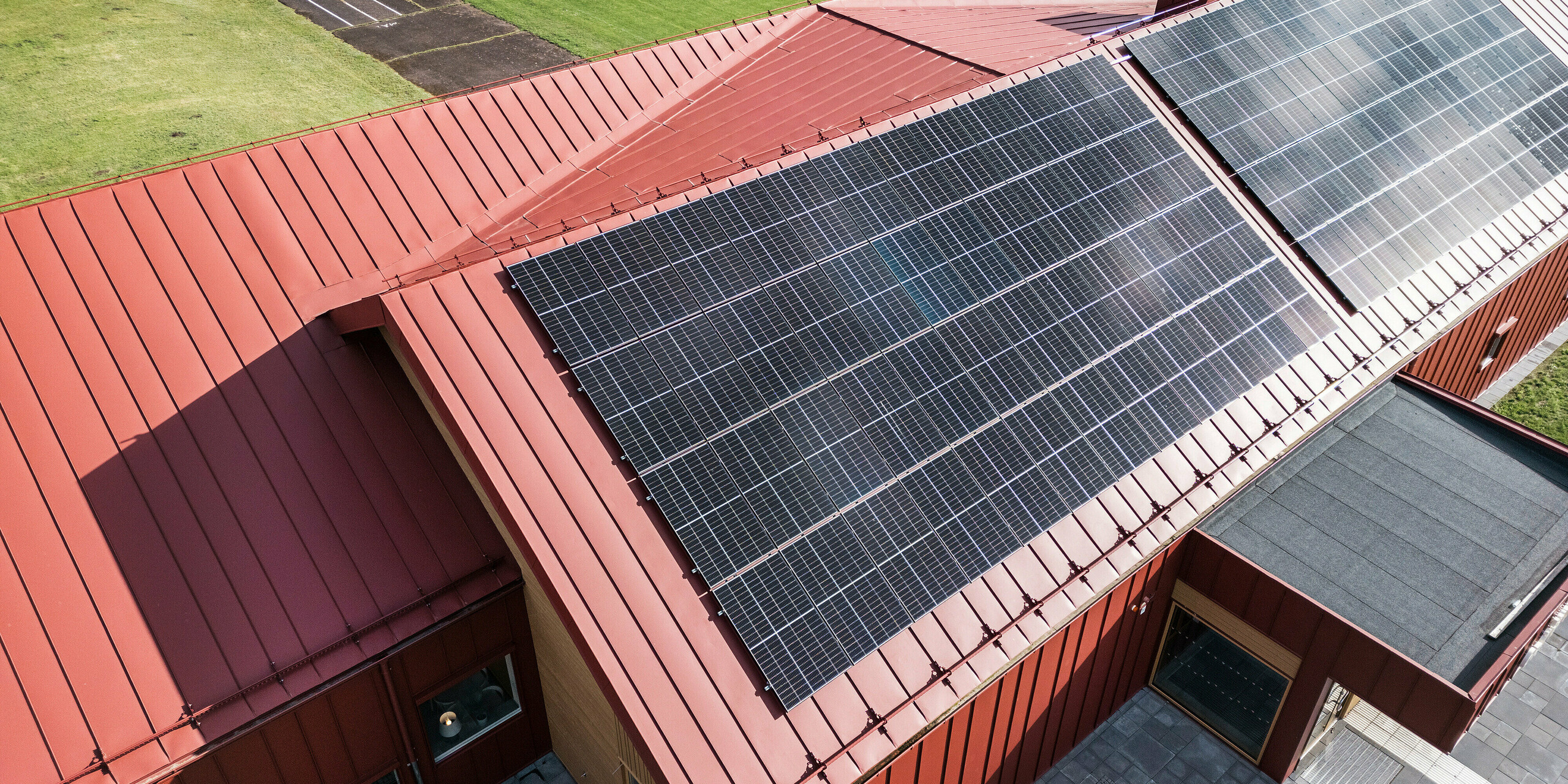 La photo aérienne de la cantine scolaire Gröna Hjärtat à Broby, en Suède, montre le toit avec le revêtement à joints debout de haute qualité en PREFALZ en rouge oxyde. Un système photovoltaïque à grande échelle fournissant de l'énergie renouvelable est visible sur le toit. Le bâtiment est un exemple d’architecture moderne avec un revêtement en aluminium durable et une conception économe en énergie. La gouttière PREFA est également conçue en rouge oxyde assortie et souligne le concept esthétique global.