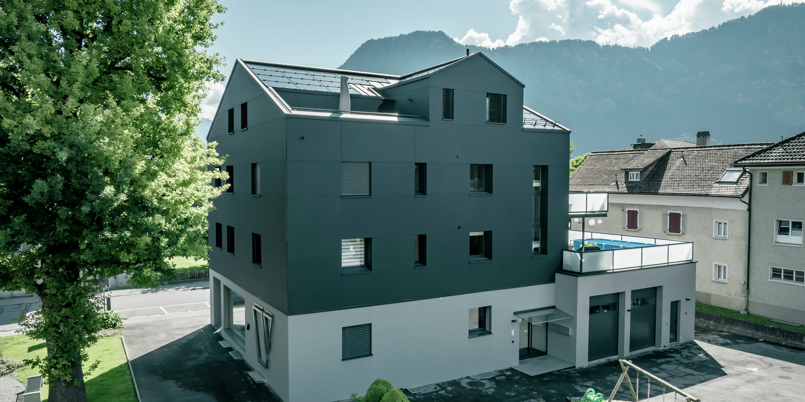 Modernisiertes Wohn- und Geschäftsgebäude mit einer anthrazitfarbenen PREFA Fassade aus PREFABOND Aluminium Verbundplatten. Die Seitenansicht zeigt klare Linien und ein zeitgemäßes Design. Im Vordergrund befinden sich gepflegte Grünflächen und ein kleiner Spielplatz. Im Hintergrund sind Berge und ein sonniger Himmel mit leichten Wolken erkennbar.