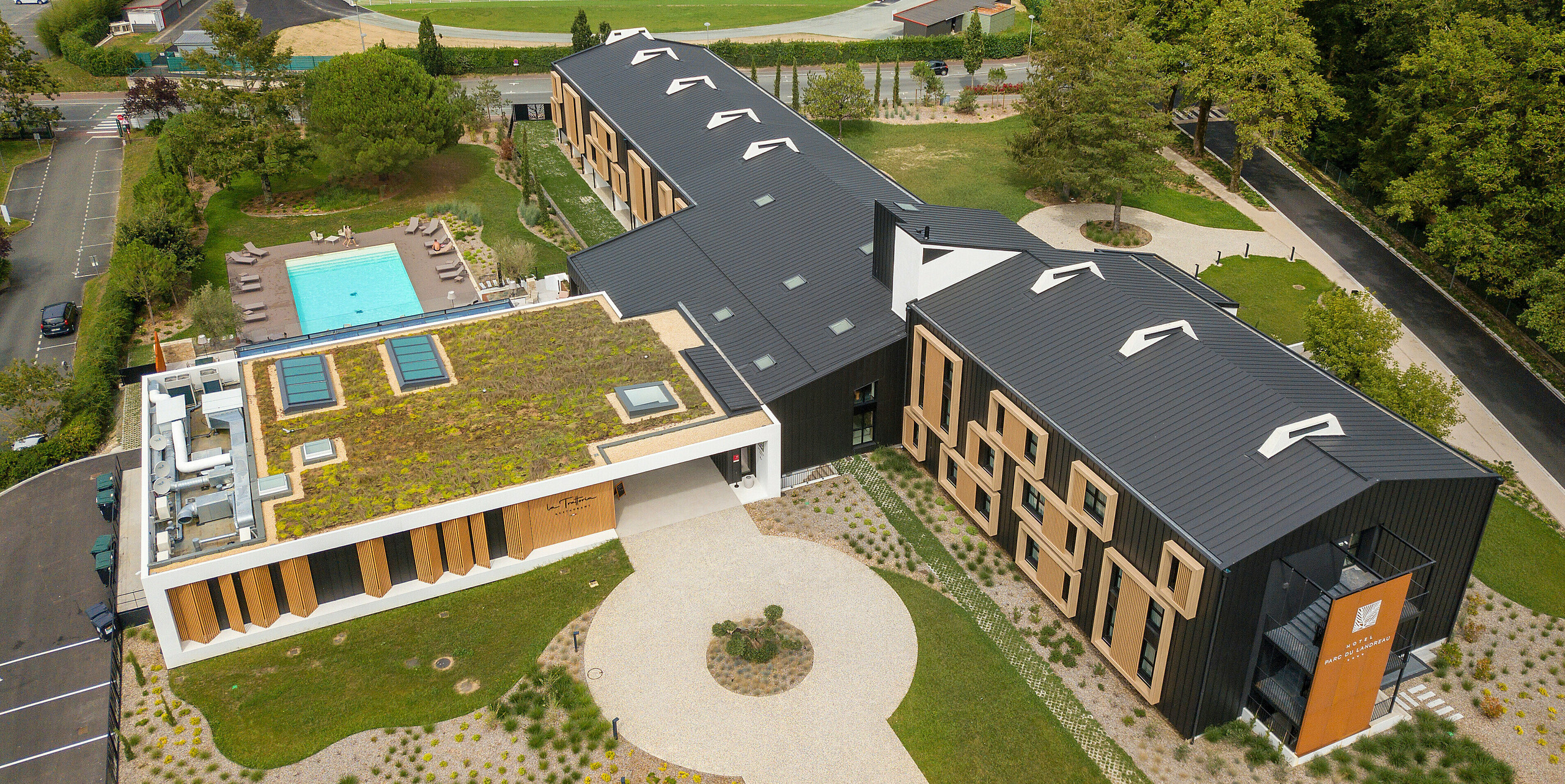 Vue aérienne de la zone d'entrée de l'Hôtel Parc du Landreau aux Herbiers, France. Le bâtiment a été revêtu du système de toiture et de façade PREFALZ en gris noir. Les cadres en bois frappants autour des fenêtres complètent le revêtement moderne en aluminium. L'espace extérieur avec piscine et toit partiellement végétalisé reflète la construction durable de l'hôtel.