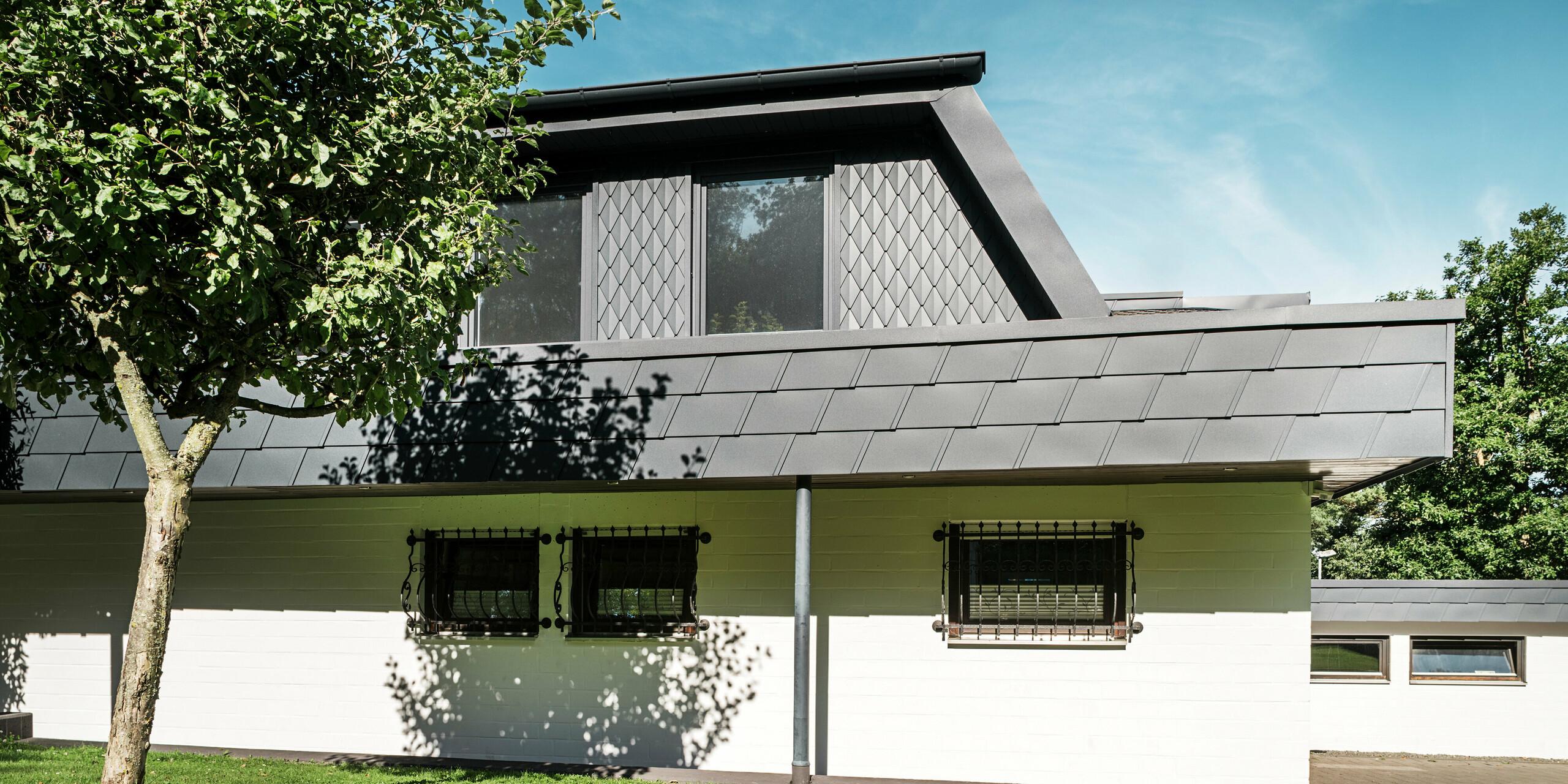 Vue latérale d'une maison individuelle à Mechernich avec une lucarne trapézoïdale impressionnante, habillée de petits losanges de toiture PREFA en P.10 anthracite. Le reste du toit est recouvert de DS.19 PREFA polyvalents de la même couleur. La façade en crépis blanc de l'étage inférieur forme un contraste élégant avec les éléments de toit sombres en aluminium robuste. Un arbre et des pelouses vertes encadrent la maison et créent un lien harmonieux avec la nature.