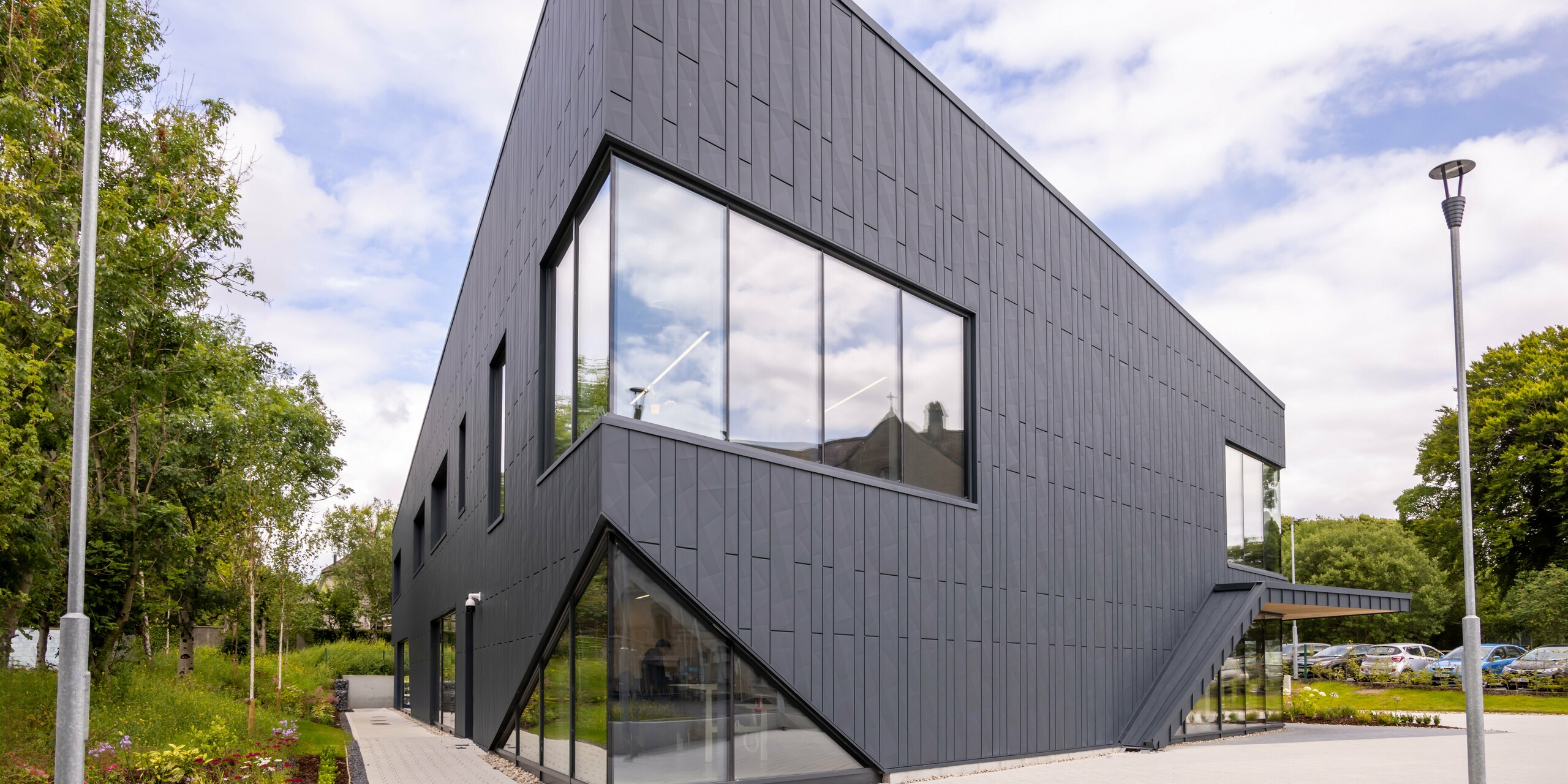 Vue de la façade du bâtiment CREW à Galway, en Irlande, avec une façade moderne composée de panneaux de façade PREFA Siding.X et PREFALZ dans la couleur P.10 anthracite. Les lignes verticales de la façade et les grandes surfaces vitrées soulignent le design innovant et dynamique du bâtiment. Le revêtement en aluminium s'intègre harmonieusement dans le paysage environnant et souligne l'architecture de haute qualité de la propriété.