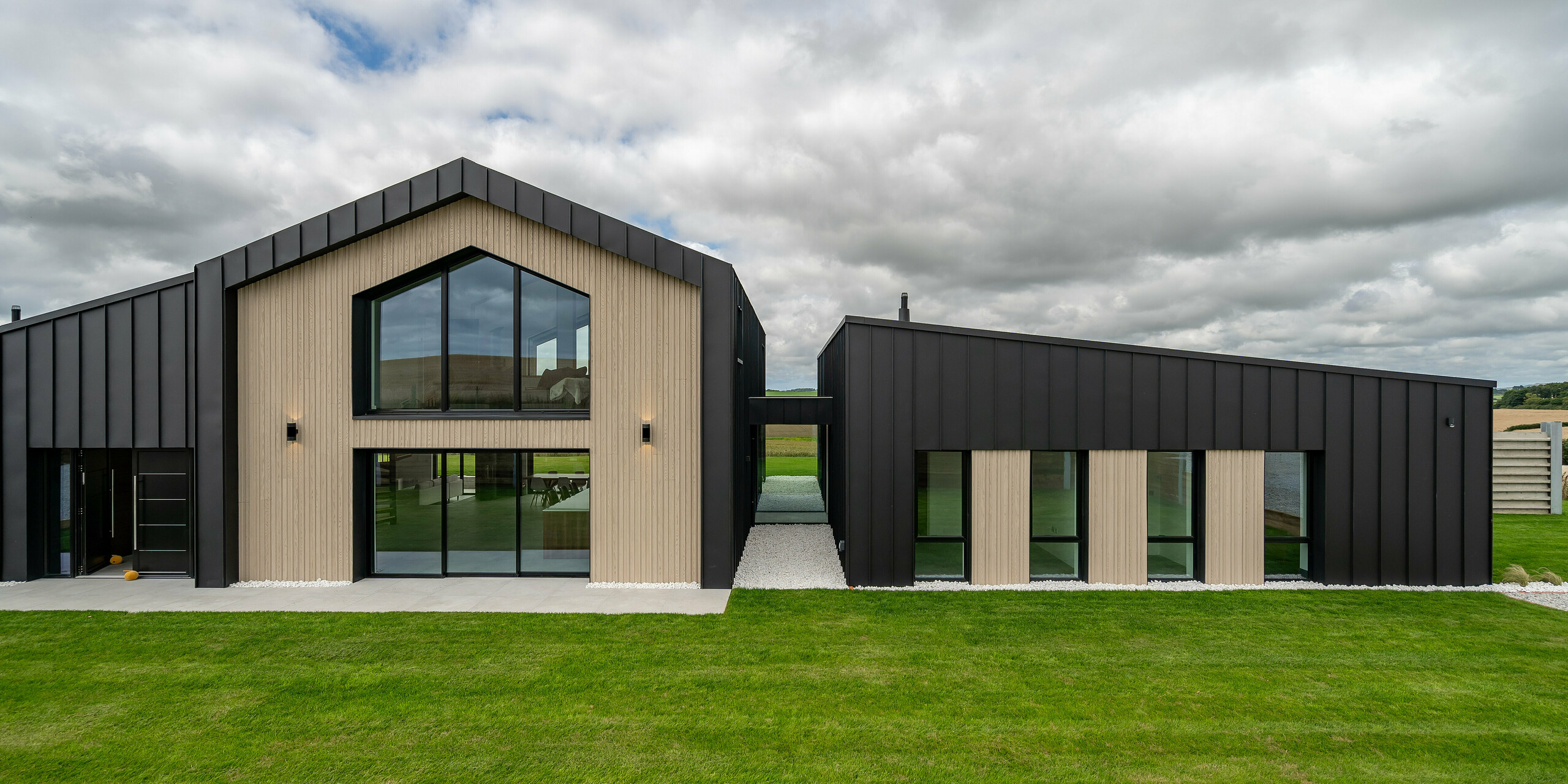 Vue de face de la maison individuelle moderne « The Hide » à Cornwall avec toiture PREFALZ et revêtement de façade en P.10 anthracite. Des lignes claires, des éléments en bois clair et de grandes surfaces vitrées complètent la toiture à joint debout foncée de PREFA. La coque robuste en aluminium confère au bâtiment une élégance intemporelle avec une protection durable contre les intempéries.