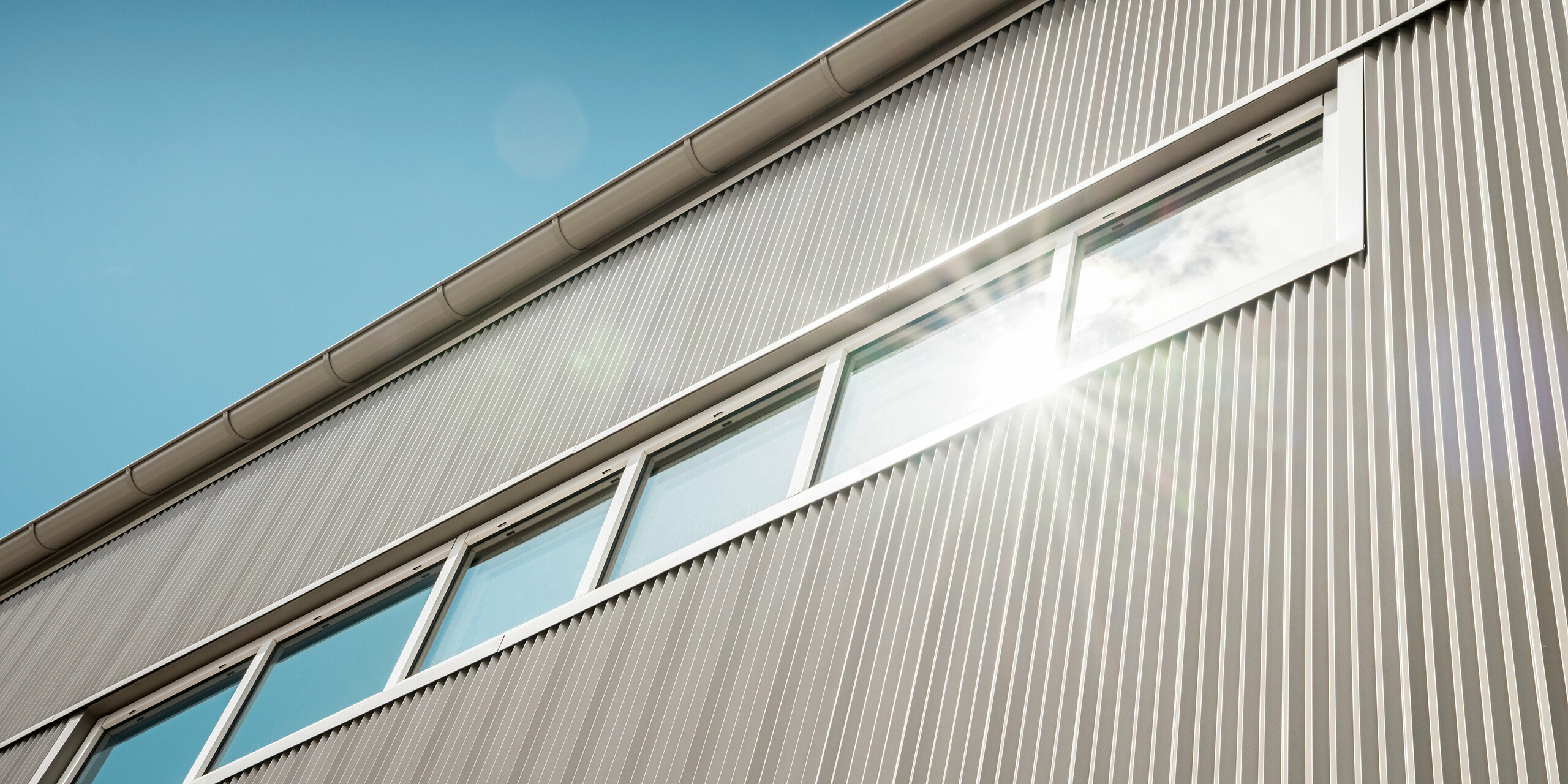 Detailansicht der PREFA Fassade aus Aluminium an der Park Arena in Värnamo, Schweden. Das PREFA Zackenprofil in Bronze erzeugt markante Licht- und Schatteneffekte. Im Bild sichtbar sind die integrierten Fenster, die perfekt in das moderne Fassadensystem eingebettet sind und das elegante Design unterstreichen. In einem Fenster spiegelt sich die Sonne.