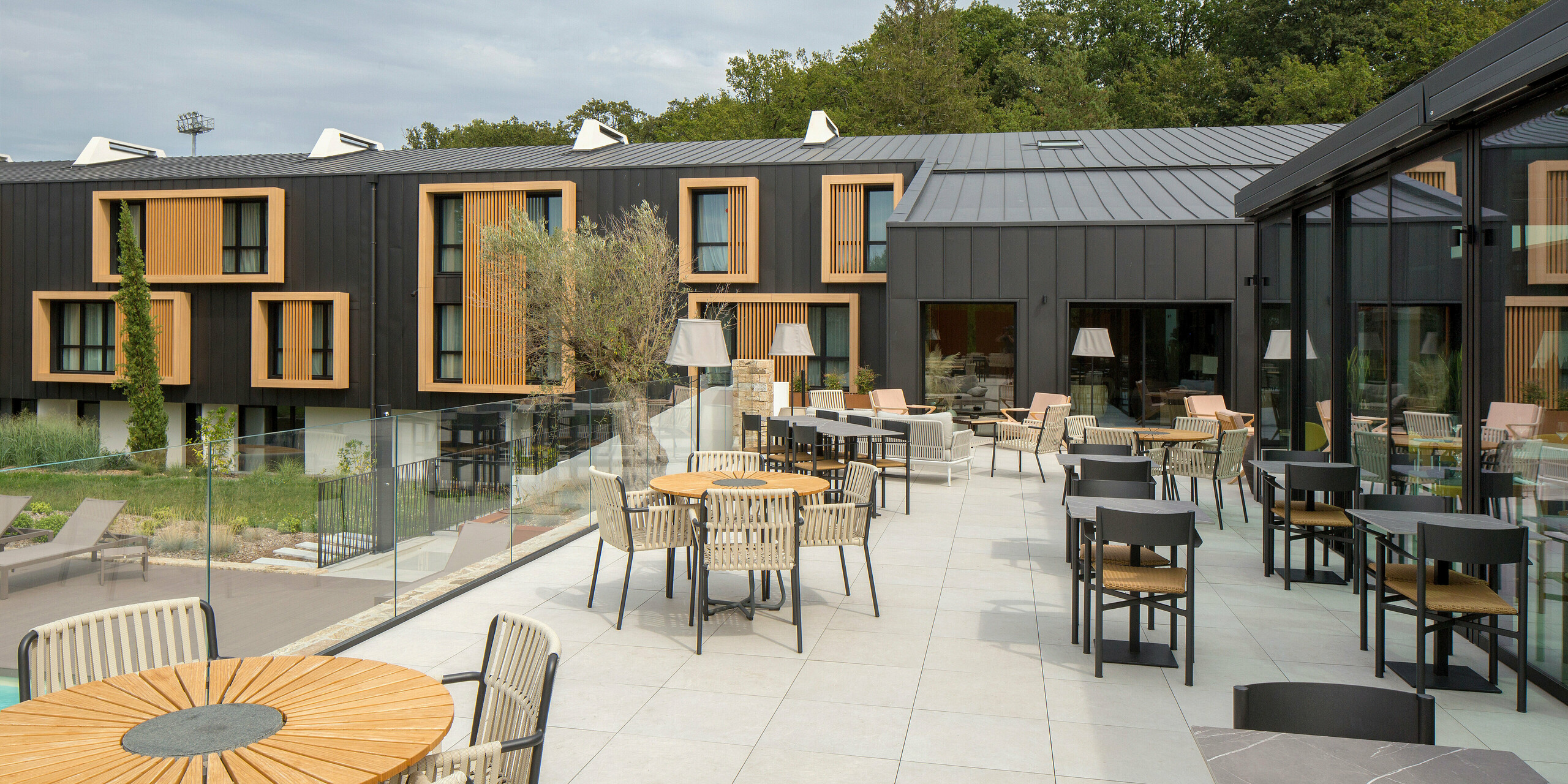 Terrasse de l'Hôtel Parc du Landreau aux Herbiers, France, avec vue sur le système de toiture et de façade PREFALZ gris noir. Les cadres en bois naturel autour des fenêtres complètent le revêtement en aluminium résistant aux intempéries. L'espace extérieur offre aux hôtes une atmosphère accueillante avec des sièges confortables.