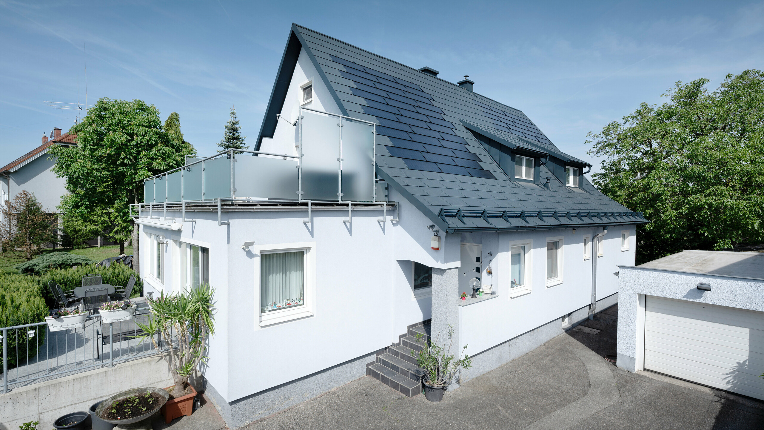 Rénovation du toit d'une maison individuelle à Leonding avec des tuiles solaires PREFA en anthracite. La photo montre la maison rénovée d'un point de vue latéral avec une vue dégagée sur le toit et les modules solaires intégrés. Ceux-ci offrent un moyen durable et économe en énergie de produire de l’électricité. Dans cette propriété, les tuiles solaires génèrent une puissance électrique de 10,6 kWc. Les matériaux PREFA modernes, durables et résistants aux intempéries contribuent à une construction de toiture esthétique et fonctionnelle qui répond aux dernières normes en matière de construction durable.