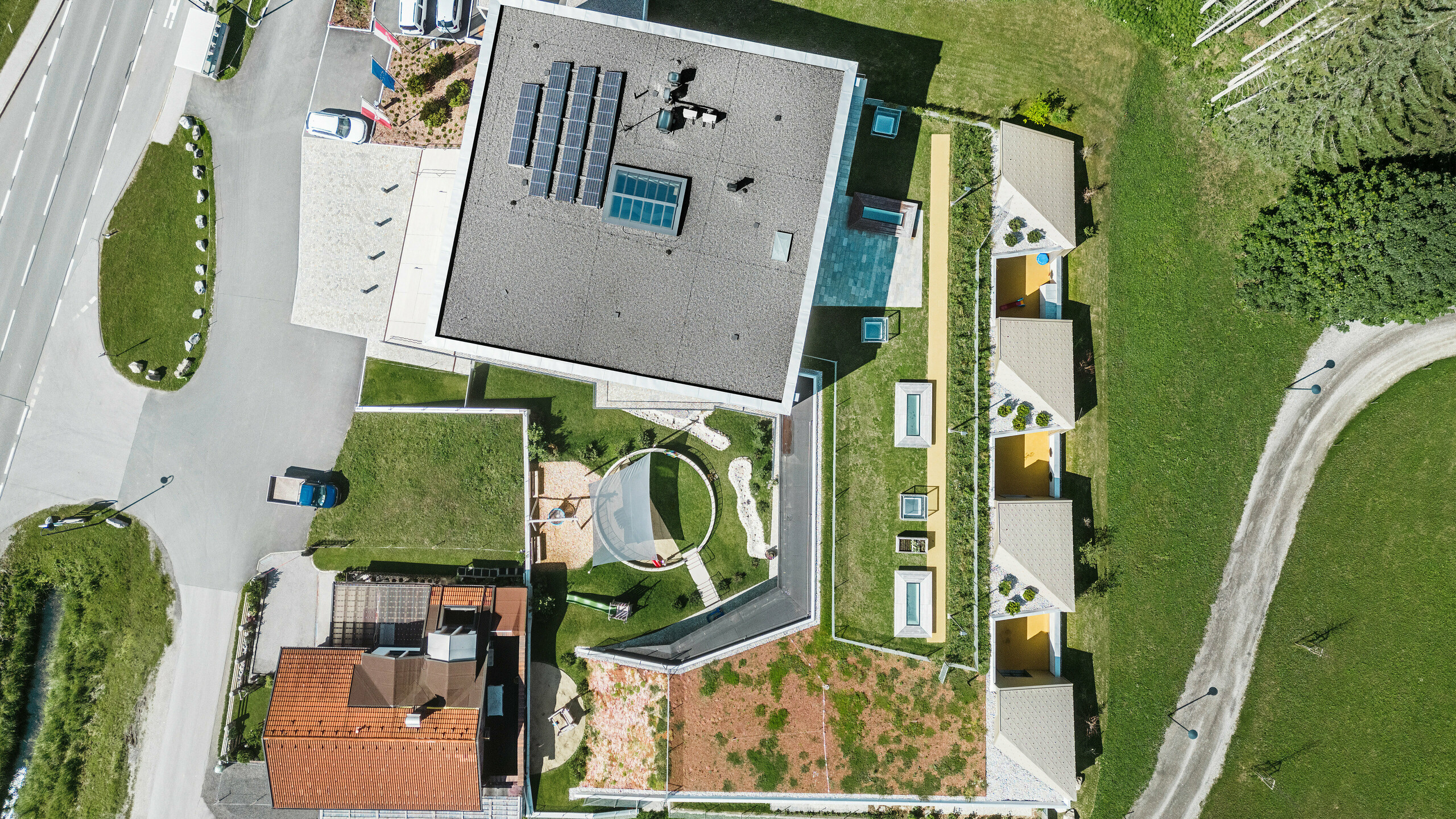 Vue aérienne du centre de formation de Gerlos, avec un ensemble de bâtiments modernes et traditionnels. Le bâtiment principal se présente avec un toit incliné gris équipé de panneaux solaires et est entouré d'autres bâtiments aux façades jaunes et aux toits verts. En arrière-plan se dessine le pittoresque paysage alpin boisé, tandis qu'au premier plan se trouvent des voitures garées et une route sinueuse.