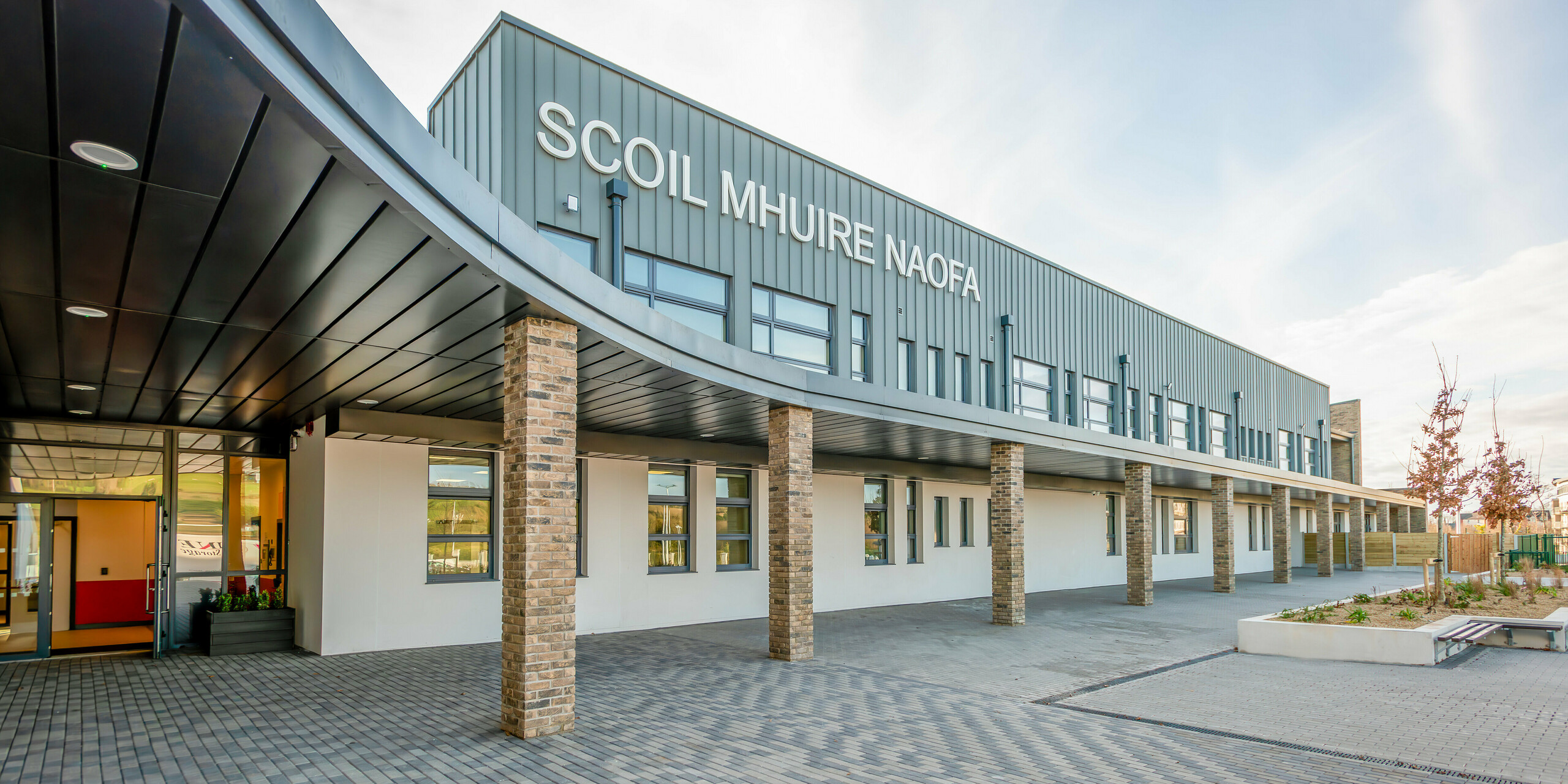 Vue détaillée de l'entrée principale du nouveau Scoil Mhuire Naofa à Carrigtwohill, en Irlande. La façade moderne en aluminium PREFALZ en P.10 gris souris associée à des colonnes en briques confère au bâtiment une élégance intemporelle. La zone couverte et les grandes fenêtres sur tout le côté du bâtiment créent une atmosphère accueillante pour les étudiants et les visiteurs.