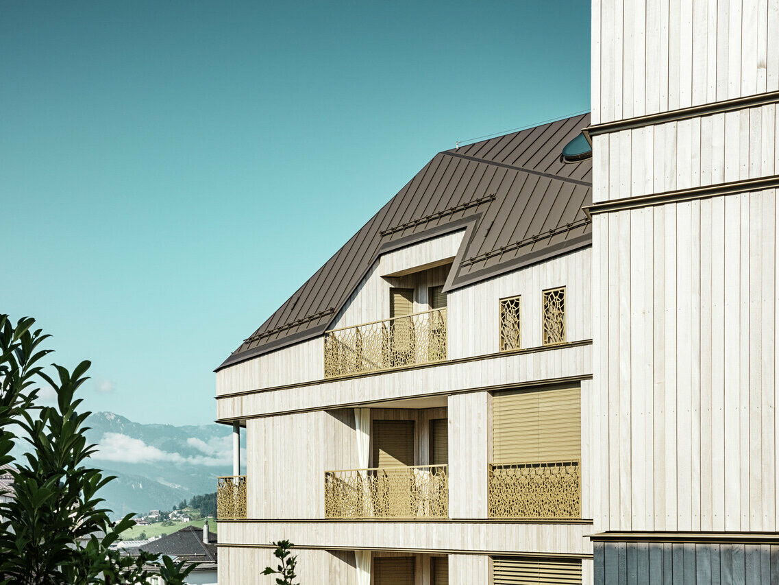 Gros plan sur un immeuble d'habitation moderne à Liebwylen, en Suisse, avec un toit en aluminium PREFALZ de couleur brune. La façade à l'aspect bois est complétée par des balustrades artistiquement conçues. En arrière-plan, des montagnes et un ciel bleu, tandis qu'une partie de la végétation environnante est visible au premier plan.