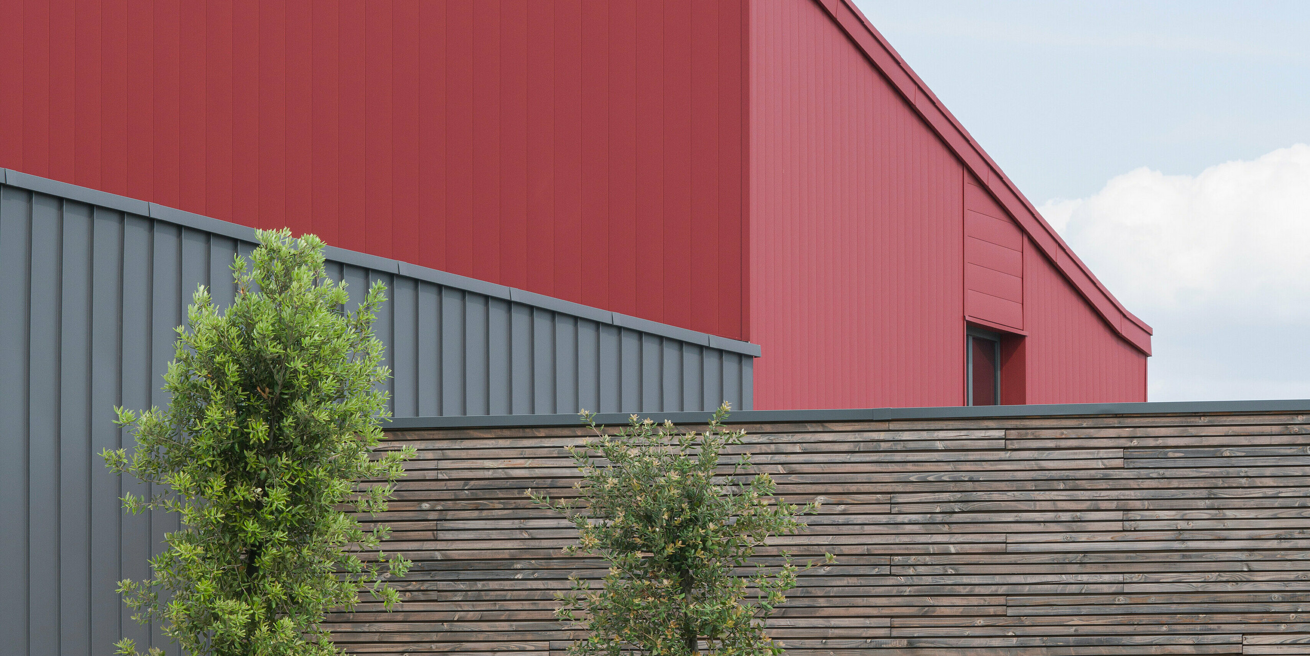 Un bâtiment commercial moderne avec une façade en aluminium rouge saisissante. Le choix de la couleur parle un langage architectural clair. Le revêtement en aluminium gris structuré forme un léger contraste avec le rouge vif. Le revêtement en aluminium PREFALZ durable sur la façade et le toit offre une protection idéale contre toutes les conditions météorologiques. Le bardage complémentaire en bois naturel apporte chaleur et naturel. Devant le bâtiment, vous pouvez voir de jeunes arbres soigneusement fixés avec des poteaux de support. Un parterre de fleurs bien entretenu ajoute de la couleur et de la vie. Une borne de recharge pour véhicules électriques au premier plan montre l'engagement en faveur de solutions énergétiques durables.