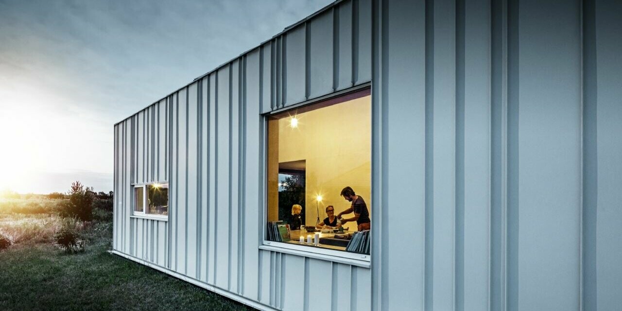 Façade avec joint debout et différentes largeurs de lames en PREFALZ de P.10 blanc PREFA photographiée au crépuscule. À travers la fenêtre de la salle à manger, on peut voir une famille en train de dîner.