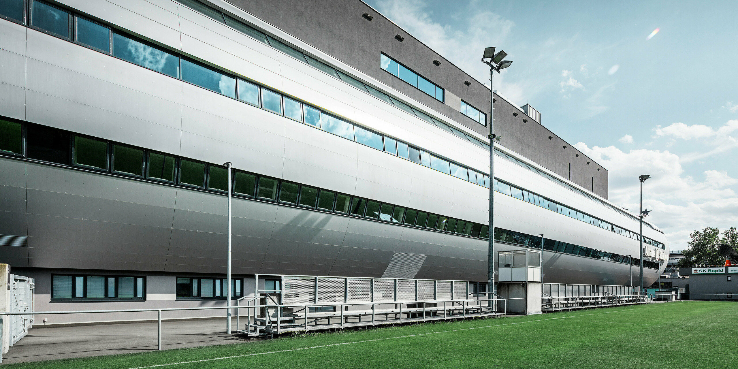 Seitenansicht des Allianz Stadions von SK Rapid Wien, charakterisiert durch die elegante PREFA Fassade in Form von Aluminium Verbundplatten in Silbermetallic. Die Architektur kombiniert Form und Funktion, mit langgezogenen Fensterfronten, die einen Blick in das sportliche Treiben gewähren und das Stadion zu einem Symbol für zeitgenössisches Design und sportliche Exzellenz machen. Der Blick vom Trainingsgelände zeigt den Blick der Spieler auf den gepflegten Rasen, die hohen Flutlichtmasten sowie die bequemen Betreuerbänke. 