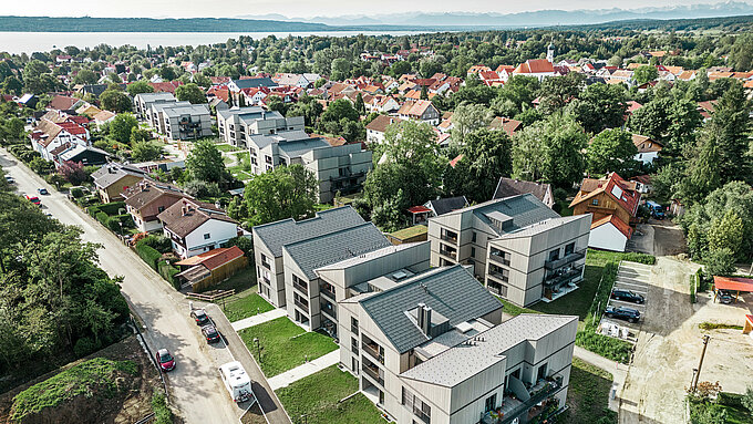 Vue panoramique du grand quartier Schmucker à Utting am Ammersee, qui se caractérise par un développement spacieux et moderne avec plusieurs immeubles d'habitation. Les six bâtiments sont équipés de R.16 PREFA durables en gris sombre et s'intègrent harmonieusement dans l'environnement. Les bâtiments sont entourés d'arbres verts et le bleu du lac Ammersee brille en arrière-plan. L'architecture de l'ensemble du projet résidentiel impressionne par ses lignes claires et sa combinaison esthétique de façades en bois et de systèmes de toiture en aluminium de haute qualité qui offrent à la fois fonctionnalité et élégance visuelle.