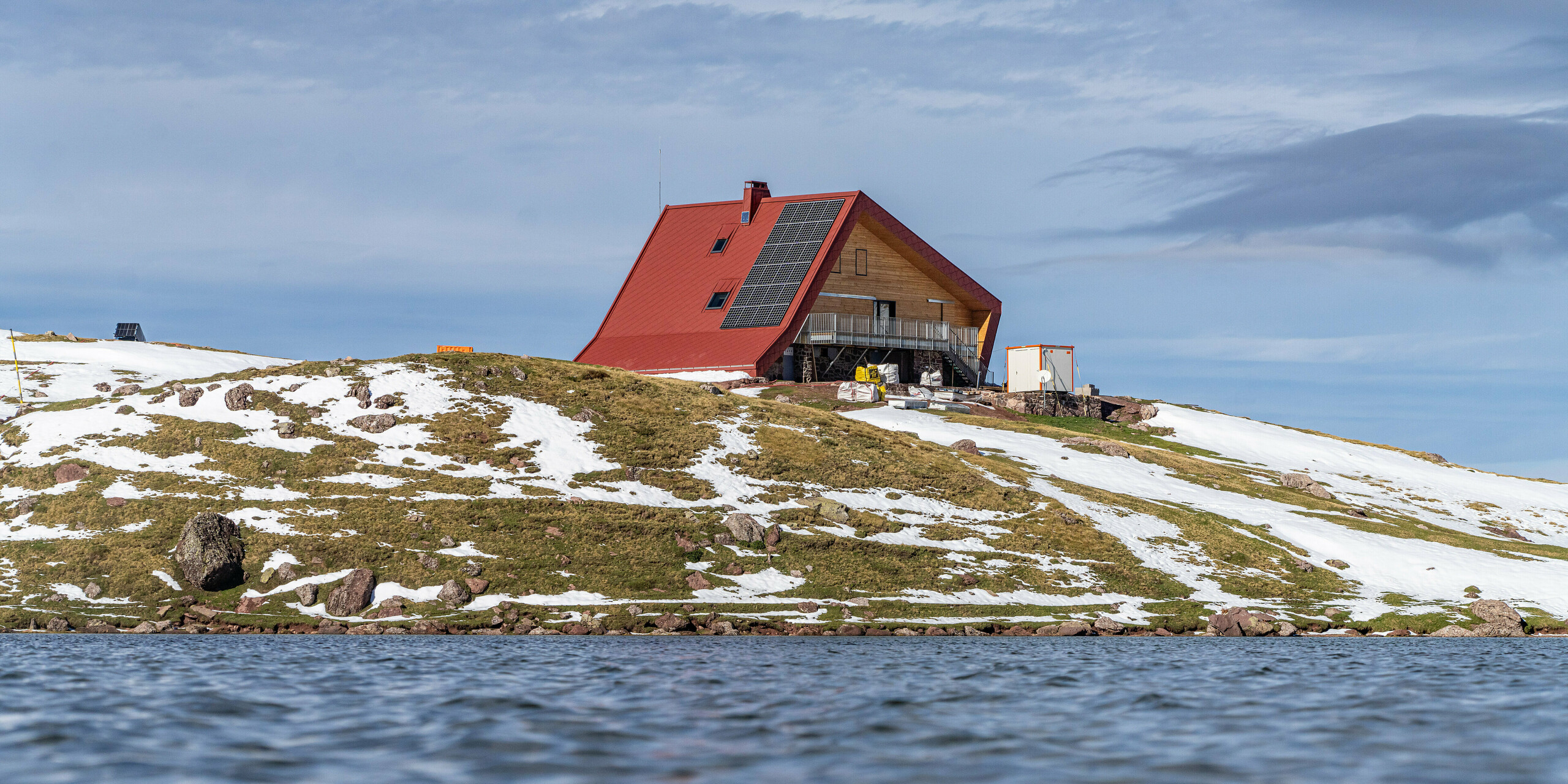 Le lac devant le refuge d'Arlet dans le Parc National des Pyrénées est au premier plan de l'image. Le bâtiment à la coque en aluminium rouge oxyde n'est visible que de loin.