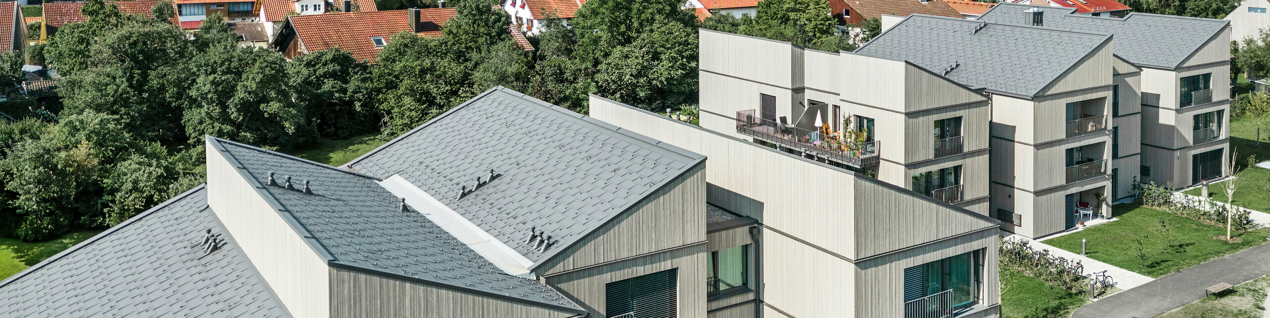 Prise de vue par drone du complexe immobilier moderne sur le site Schmucker à Utting am Ammersee (Allemagne), avec une construction hybride en bois innovante et des R.16 PREFA durables en gris sombre P.10. Les bâtiments se caractérisent par leurs lignes claires et leur architecture durable, combinées à d'élégantes façades en bois et à des balcons spacieux. La finition de haute qualité des produits en aluminium PREFA garantit une résistance aux intempéries et une longévité optimales, tandis que l'intégration harmonieuse dans l'environnement vert crée une atmosphère de vie accueillante. Au premier plan, vous pouvez voir une aire de jeux verte, qui contribue à l'aménagement familial du quartier.